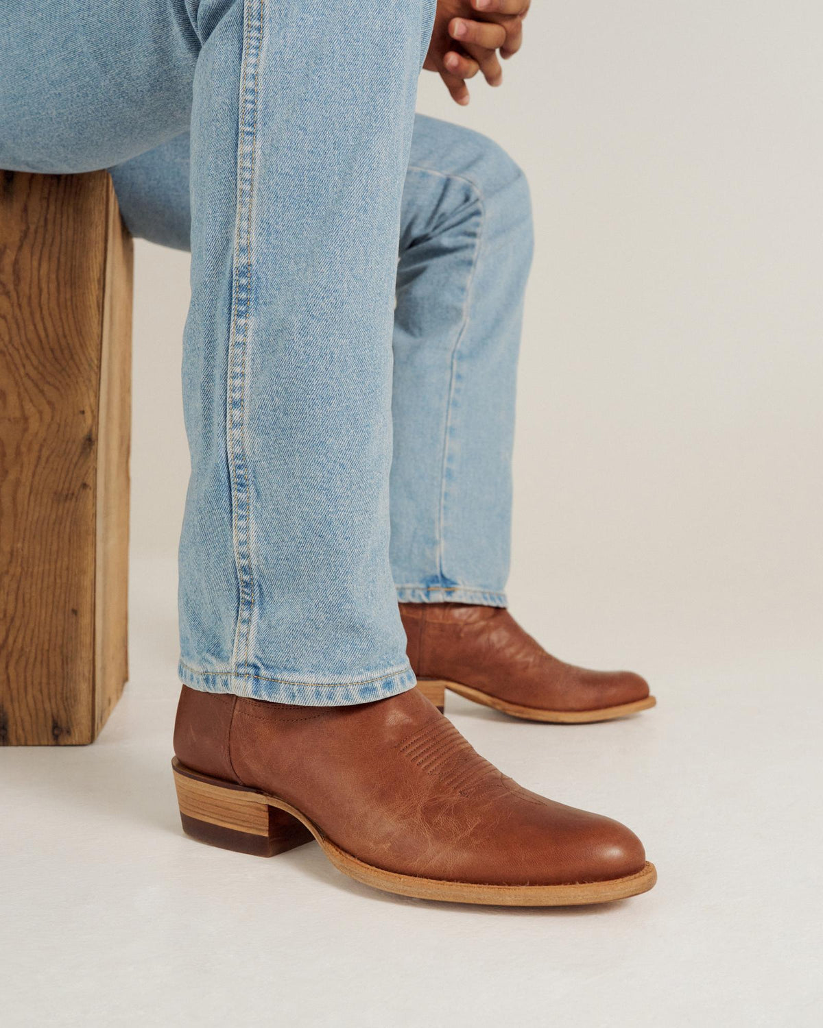Person wearing light blue jeans and brown leather heeled boots sits on a wooden stool against a plain background.