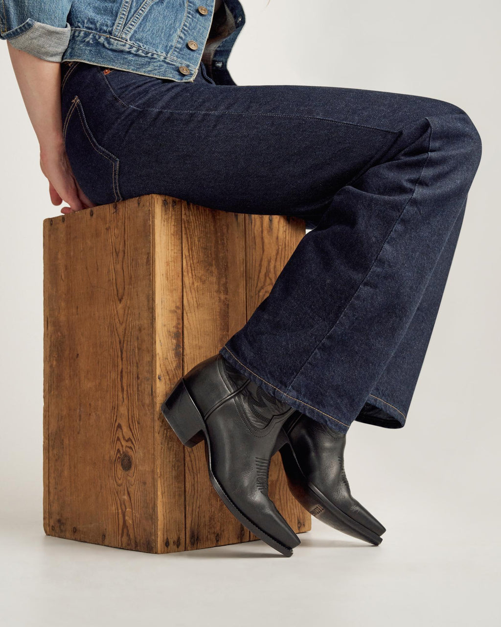 Person wearing dark blue jeans and black leather cowboy boots sits on a wooden box, with a light background visible.