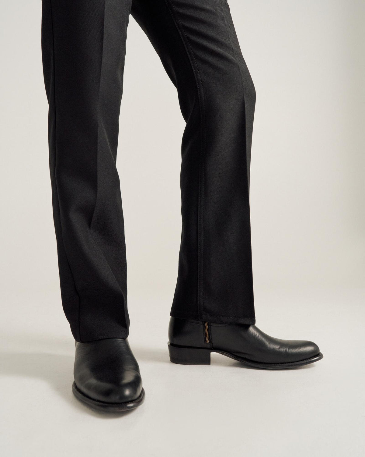 A person wearing black dress pants and black leather shoes stands on a light-colored surface.