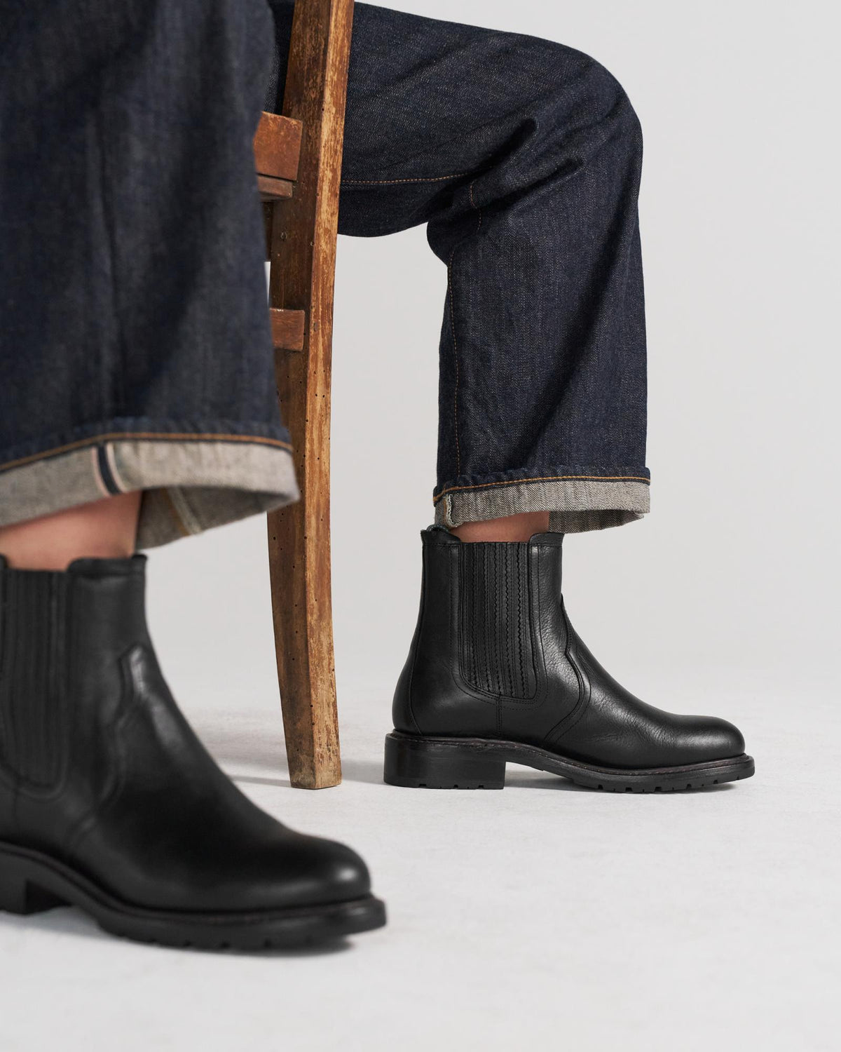 Person wearing dark rolled-up jeans and black leather ankle boots, reminiscent of The Chelsea Cowhide: Midnight, sits on a wooden chair against a plain background.