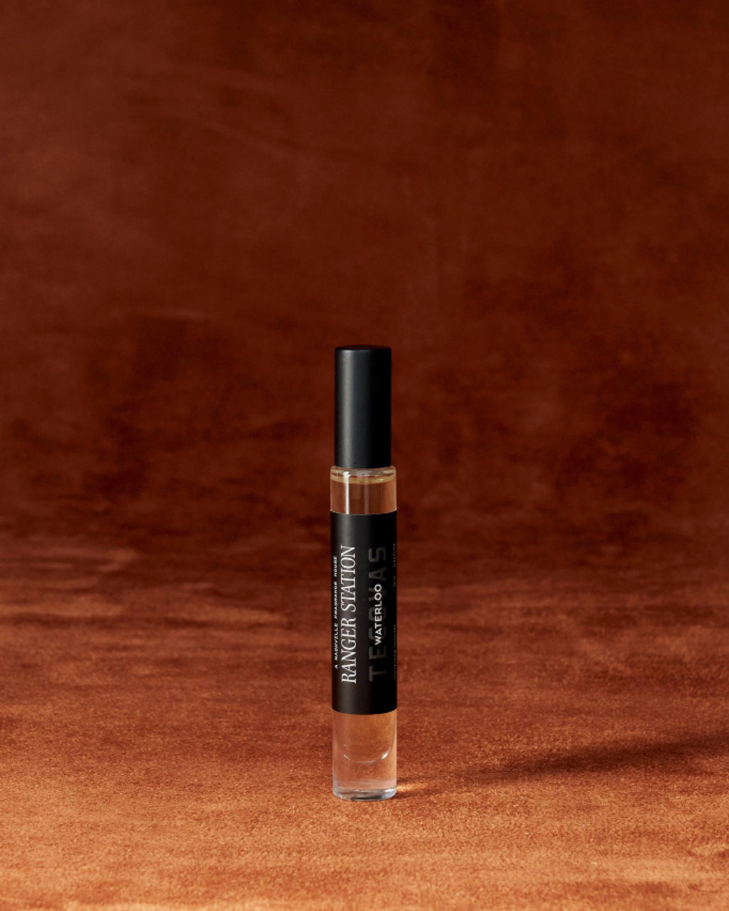 A small glass perfume bottle with a black cap and label stands upright on a brown textured surface with a matching brown background.