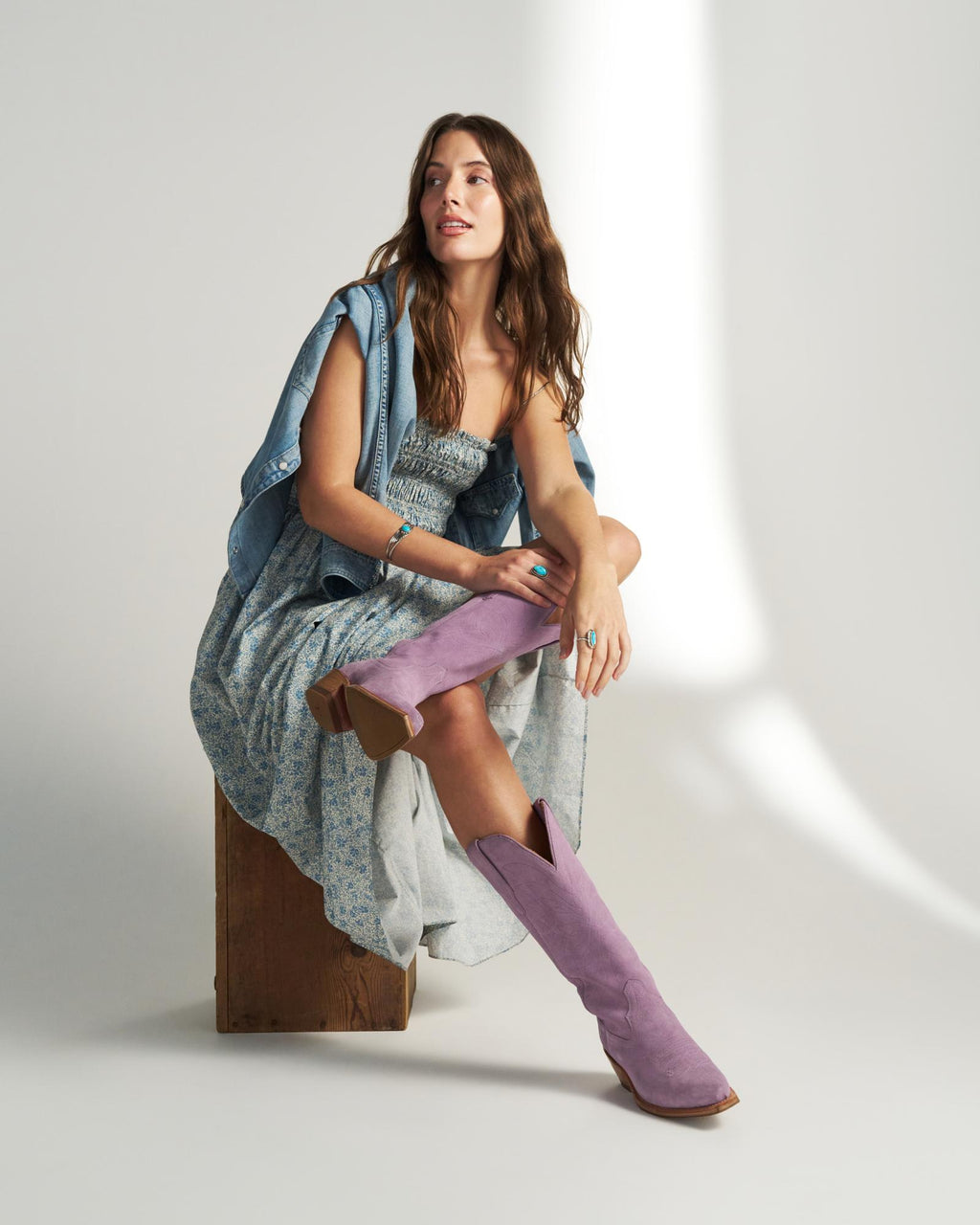 Woman sitting on a wooden block, wearing a blue floral dress, denim shirt draped over her shoulders, and tall lavender The Annie Suede boots, set against a neutral background.