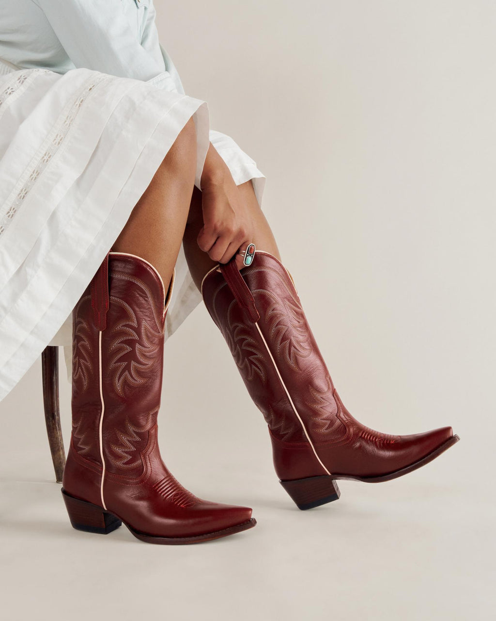 A person wearing a white skirt and red cowboy boots sits on a chair, with their hands resting on their legs against a plain background.