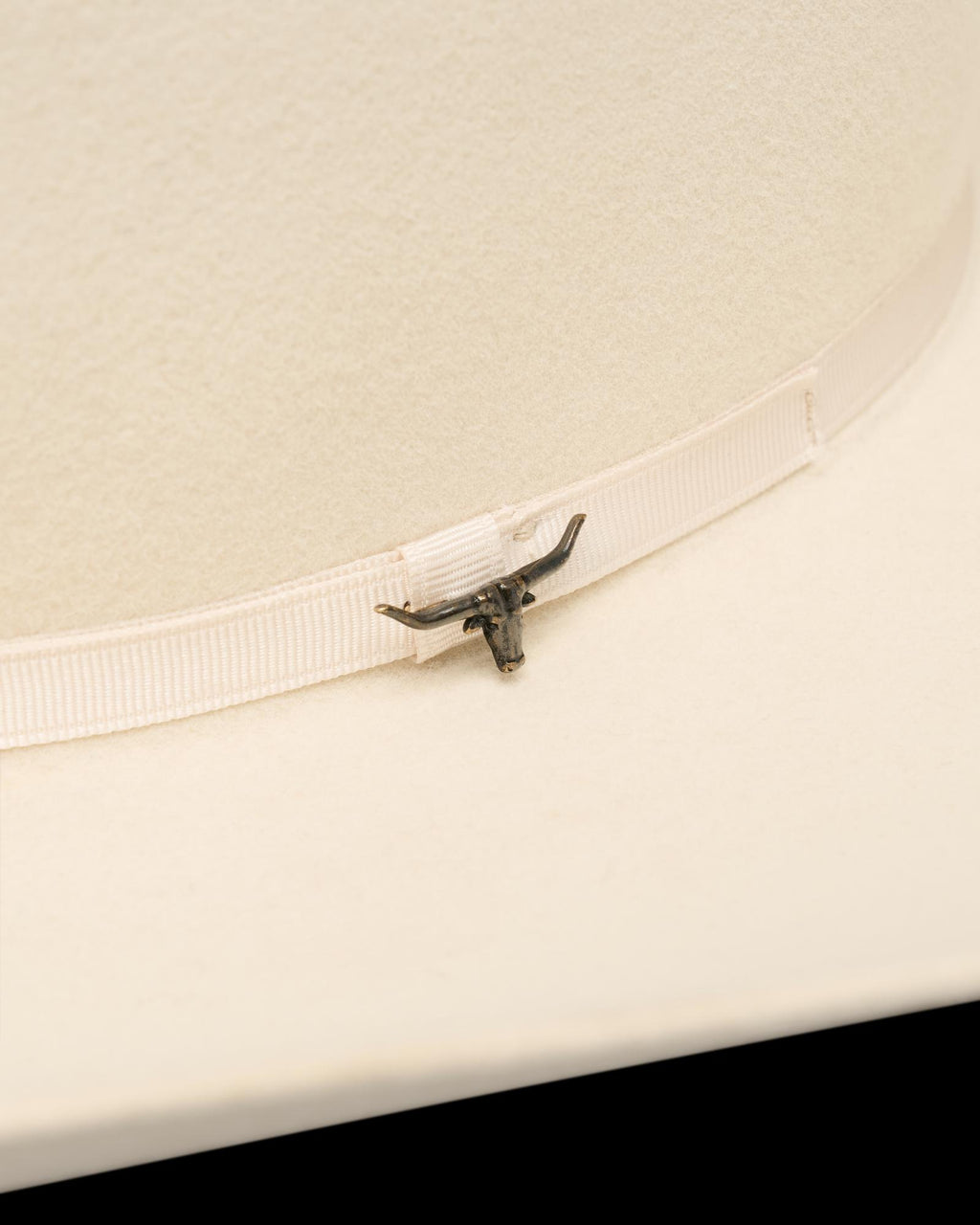 Close-up of a light-colored cowboy hat featuring a ribbon band with a small metallic longhorn emblem attached.