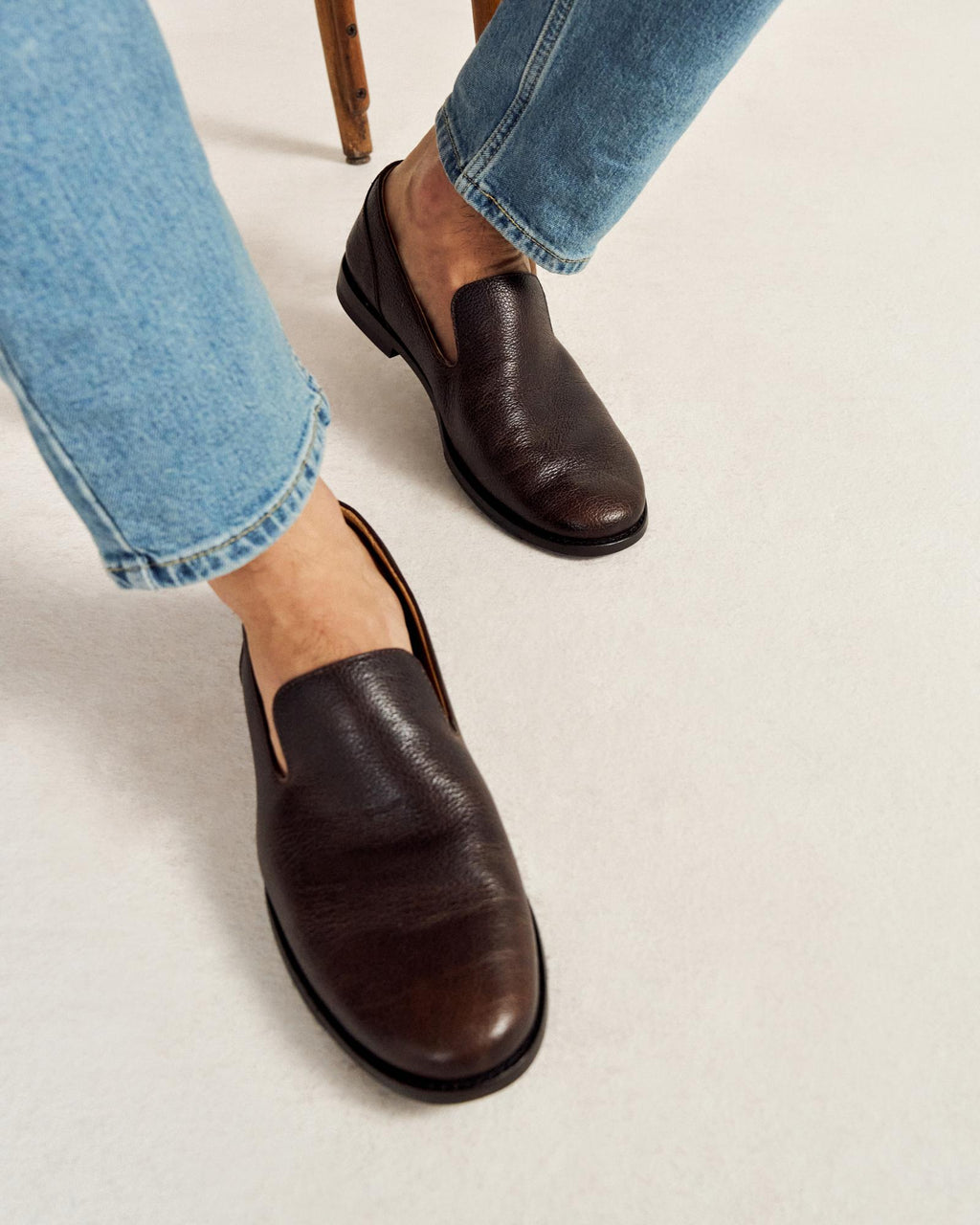 Person wearing light blue jeans and dark brown leather slip-on shoes, sitting on a light-colored surface with part of a wooden chair visible.