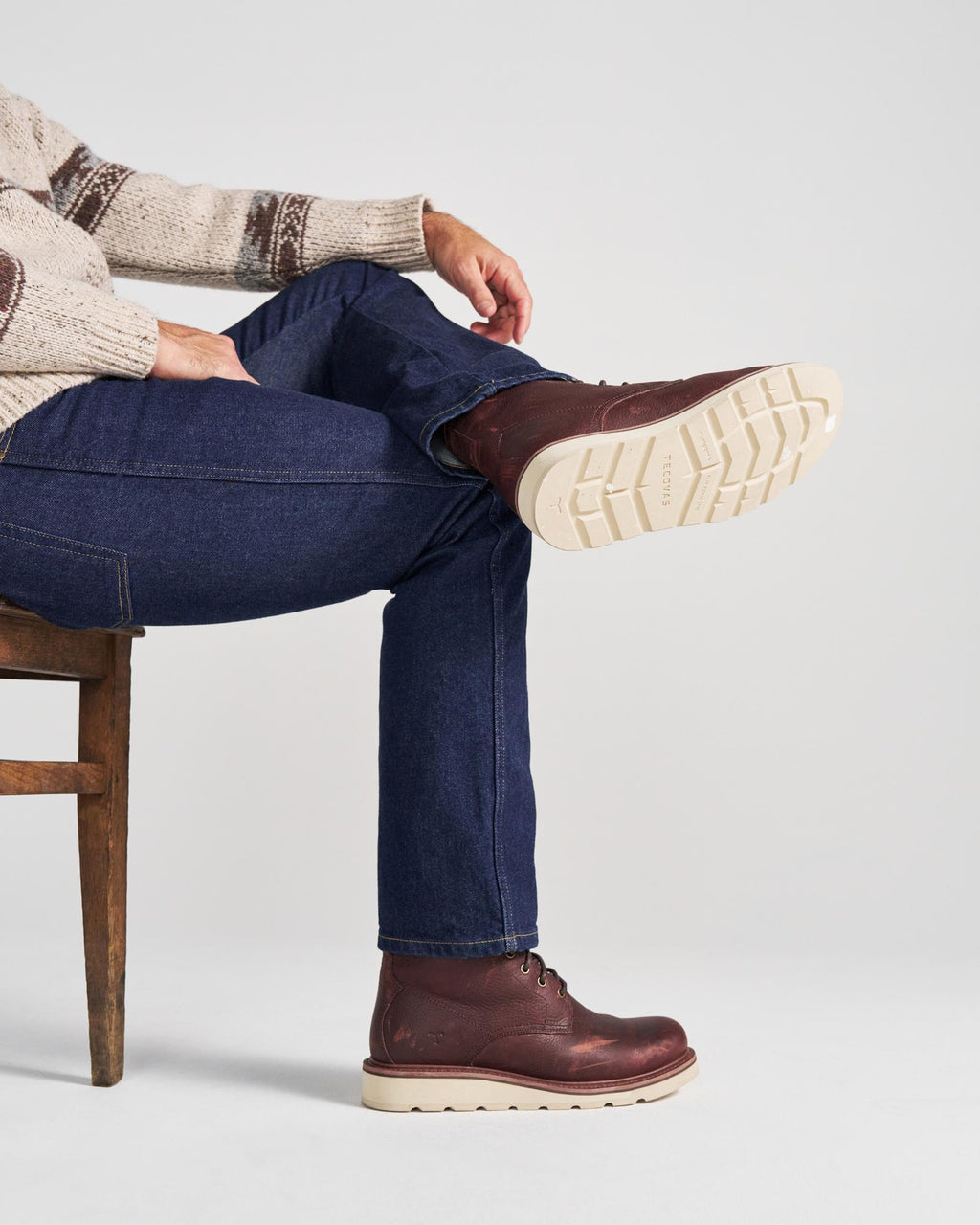 The Knox Lace Up in Cognac Cowhide. A person in jeans and a patterned sweater sits on a wooden chair, showcasing the lugged rubber soles of their Cognac Cowhide boots.
