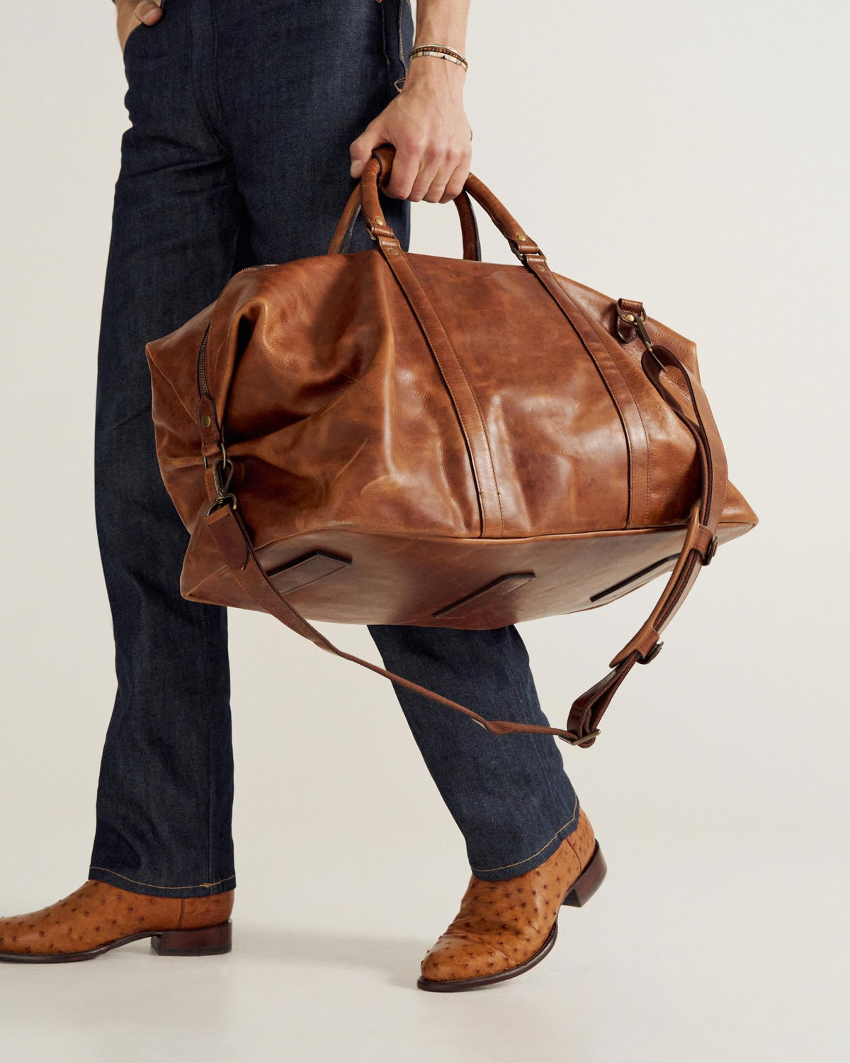 A person wearing dark jeans and tan boots holds a large brown leather duffel bag with handles and a shoulder strap.