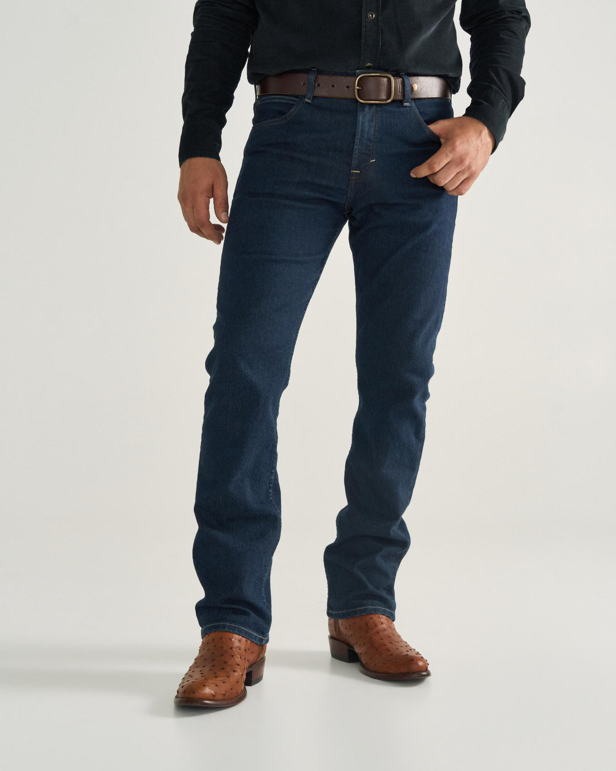 The Dean in Pecan Ostrich. A man wearing a black shirt, brown belt, dark blue jeans, and pecan ostrich cowboy boots stands against a plain background.