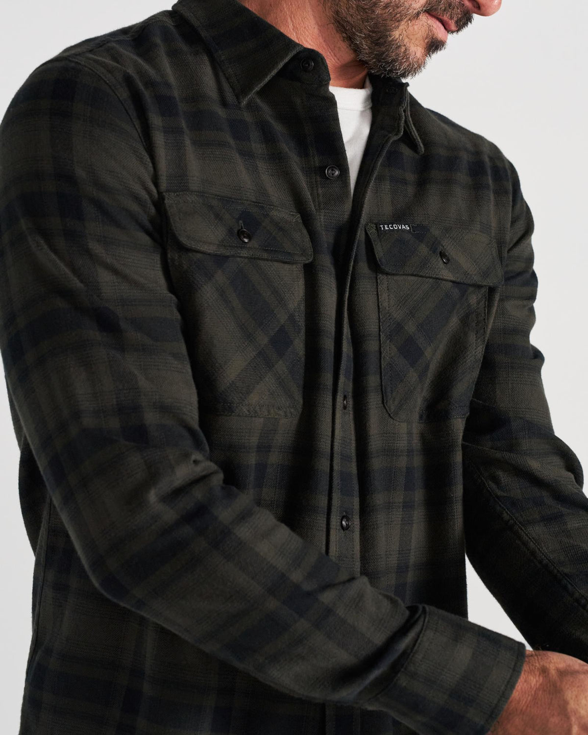 A man wearing a dark plaid button-up shirt over a white T-shirt, with the focus on the shirt’s details and the man’s hands adjusting his sleeve.