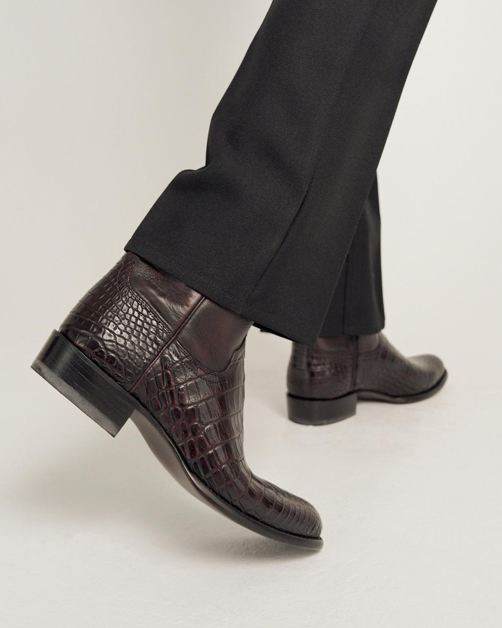 A person wearing black pants and dark brown crocodile-patterned leather ankle boots taking a step forward on a light-colored surface.