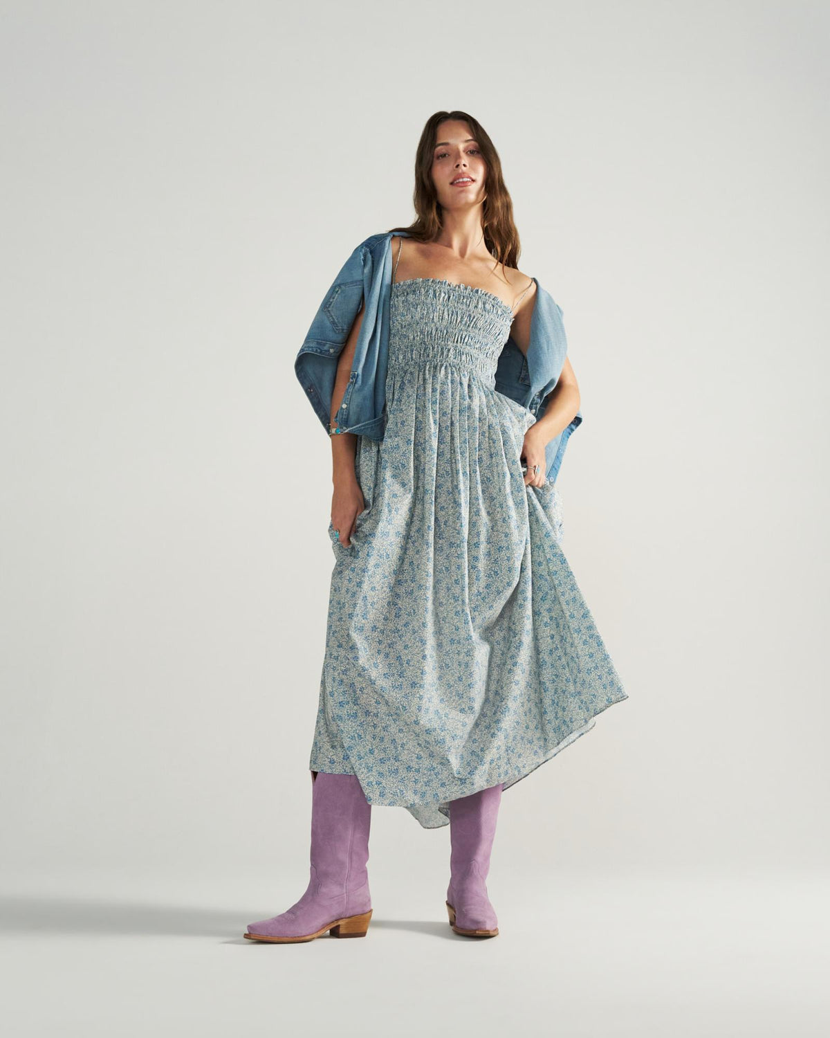 The Annie in Lavender Suede. A woman wearing a blue floral dress, a denim jacket draped over her shoulders, and Lavender Suede boots stands against a plain light background.