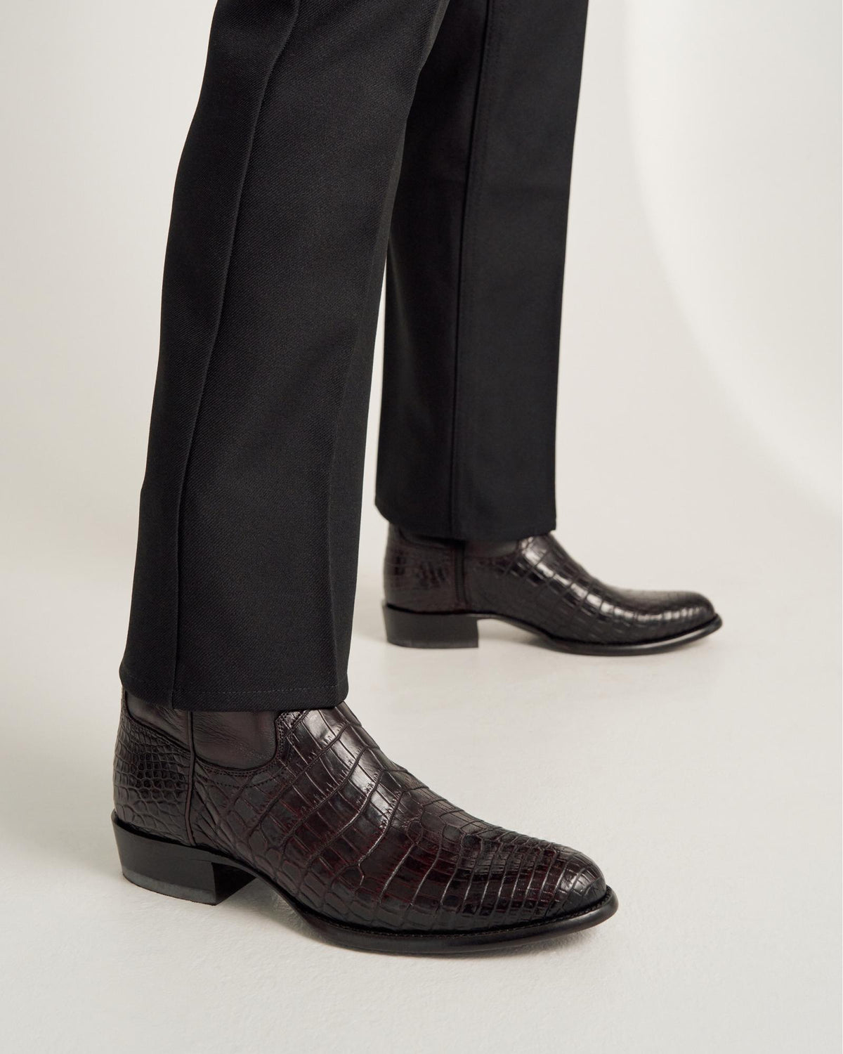 Person wearing black dress pants and dark brown, crocodile-patterned leather ankle boots standing on a white surface.