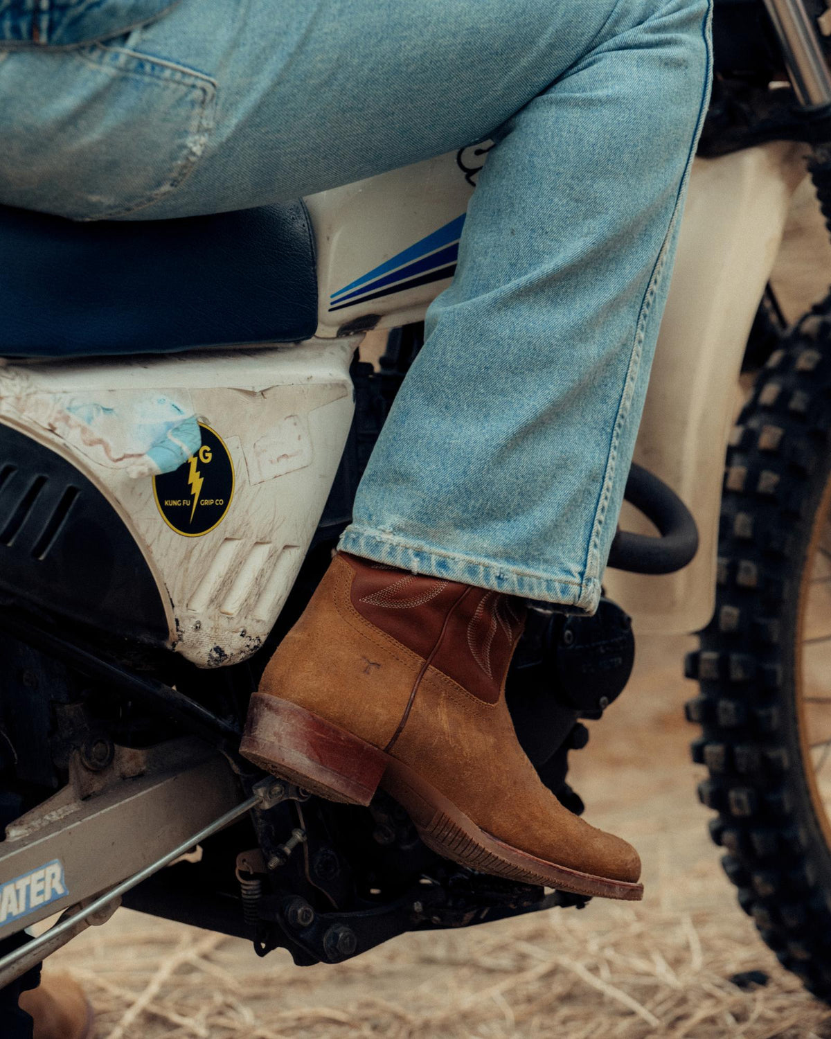 The Buck in Copper Cowhide. Person wearing jeans and brown cowhide boots sits on a white dirt bike, one foot resting on the footpeg, with dry grass scattered across the ground.