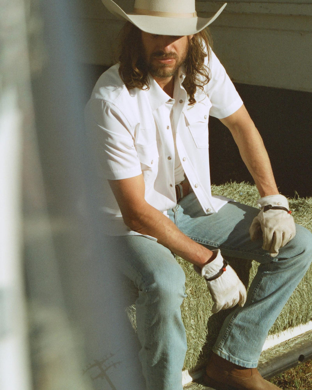 A man wearing a cowboy hat, white short-sleeve shirt, jeans, work gloves, and boots sits outdoors on a step in sunlight.