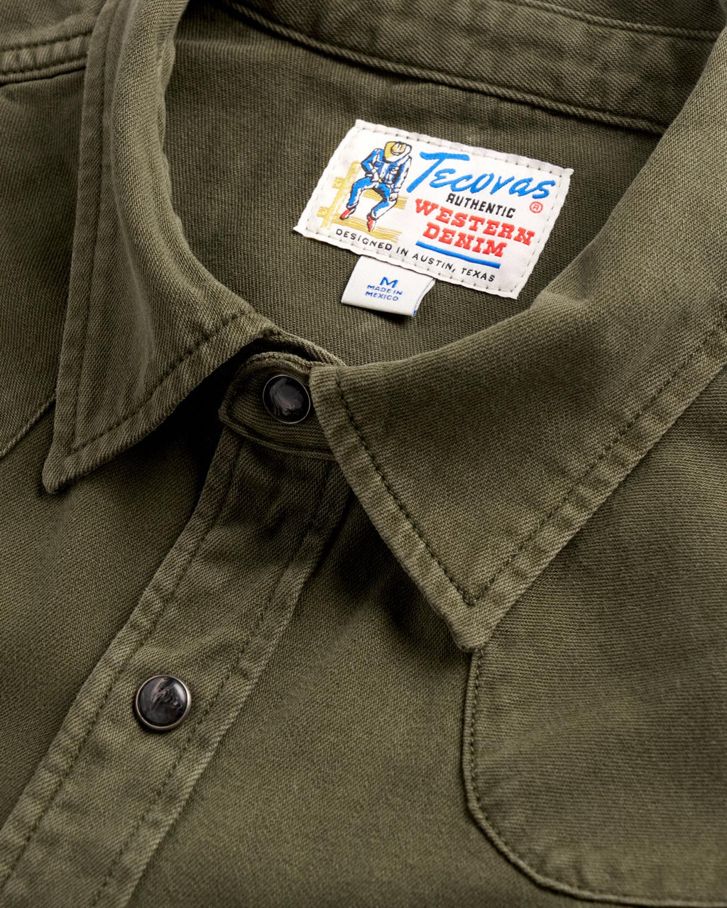 Close-up of an olive green denim shirt collar with a "Tecovas" brand label and a size M tag made in Mexico.
