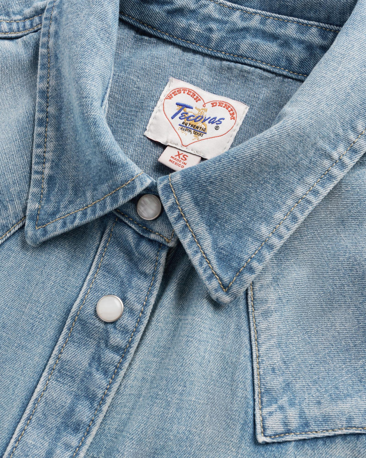 Close-up of a light blue denim shirt with metal snap buttons and a visible brand label reading “Pecos West Western Design.”.