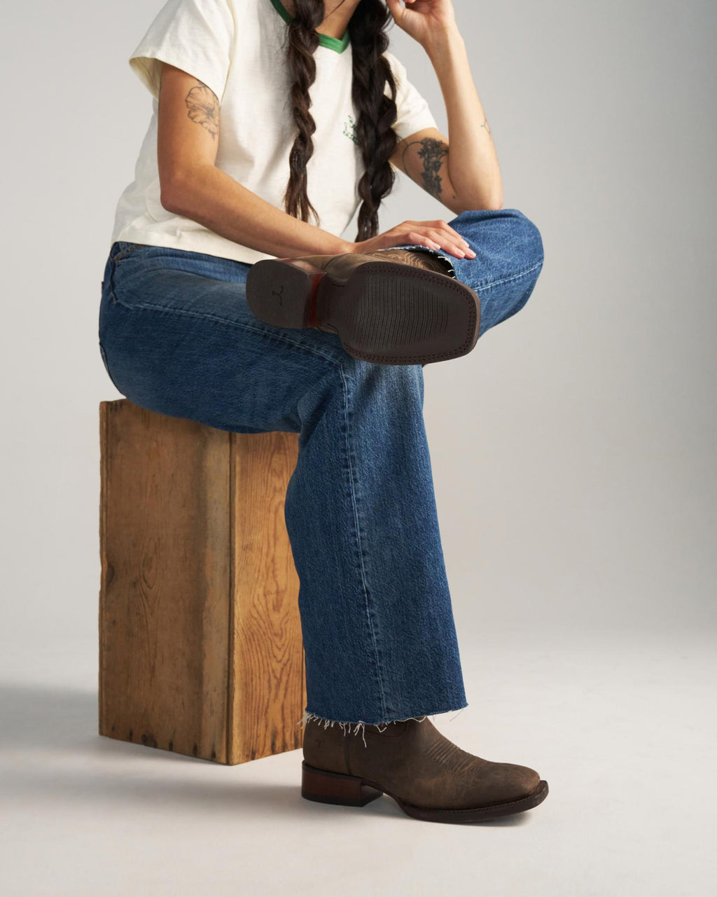The Tess in Cafe Goat. A person sits on a wooden block at Cafe Goat with one leg crossed, wearing a white t-shirt, blue jeans, and brown boots. Their face is partially out of frame.