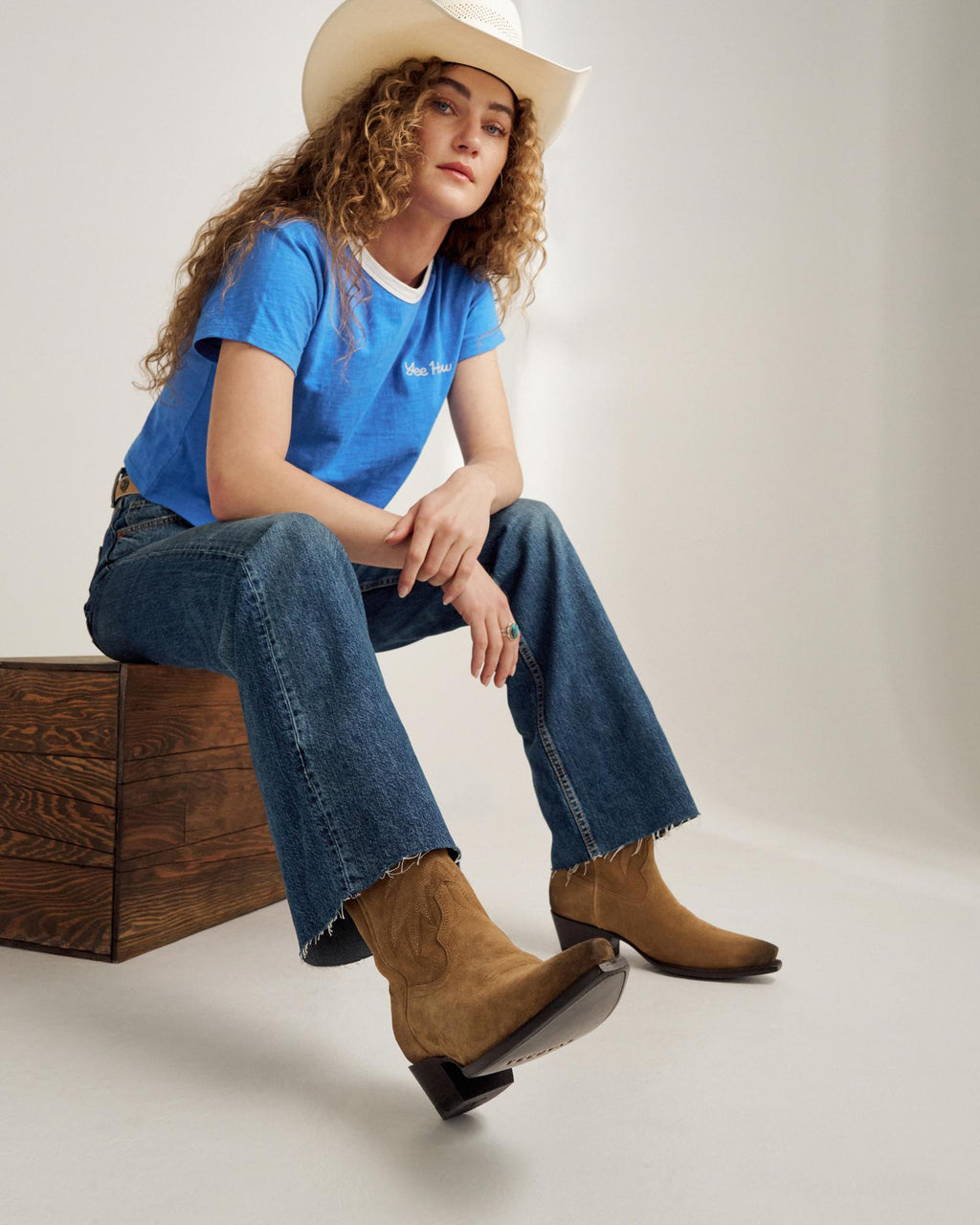 A person wearing a white cowboy hat, blue t-shirt, jeans, and tan cowboy boots sits on a wooden block against a plain background.