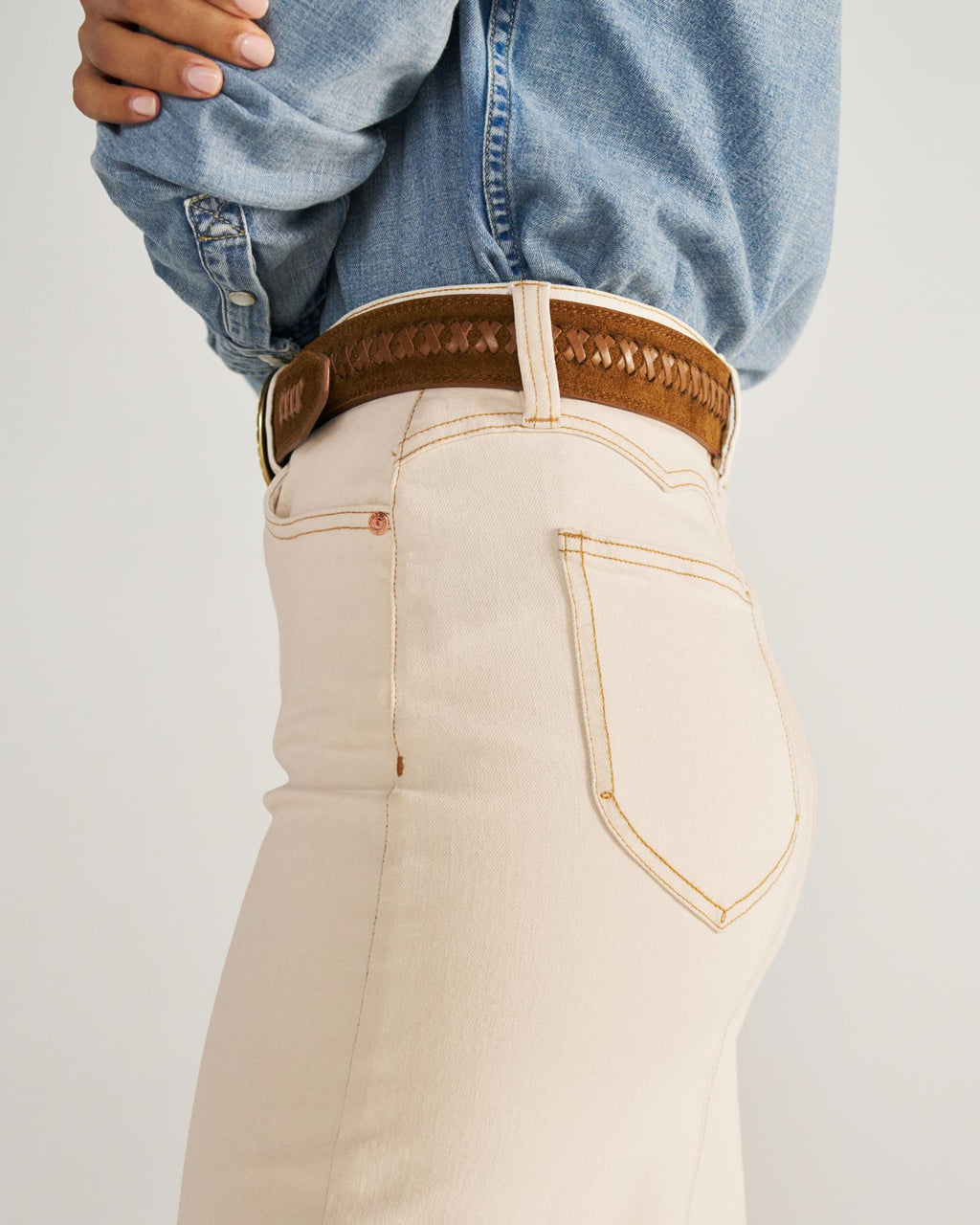 Person wearing a light blue denim shirt tucked into high-waisted beige jeans with a brown woven belt, shown from the side.