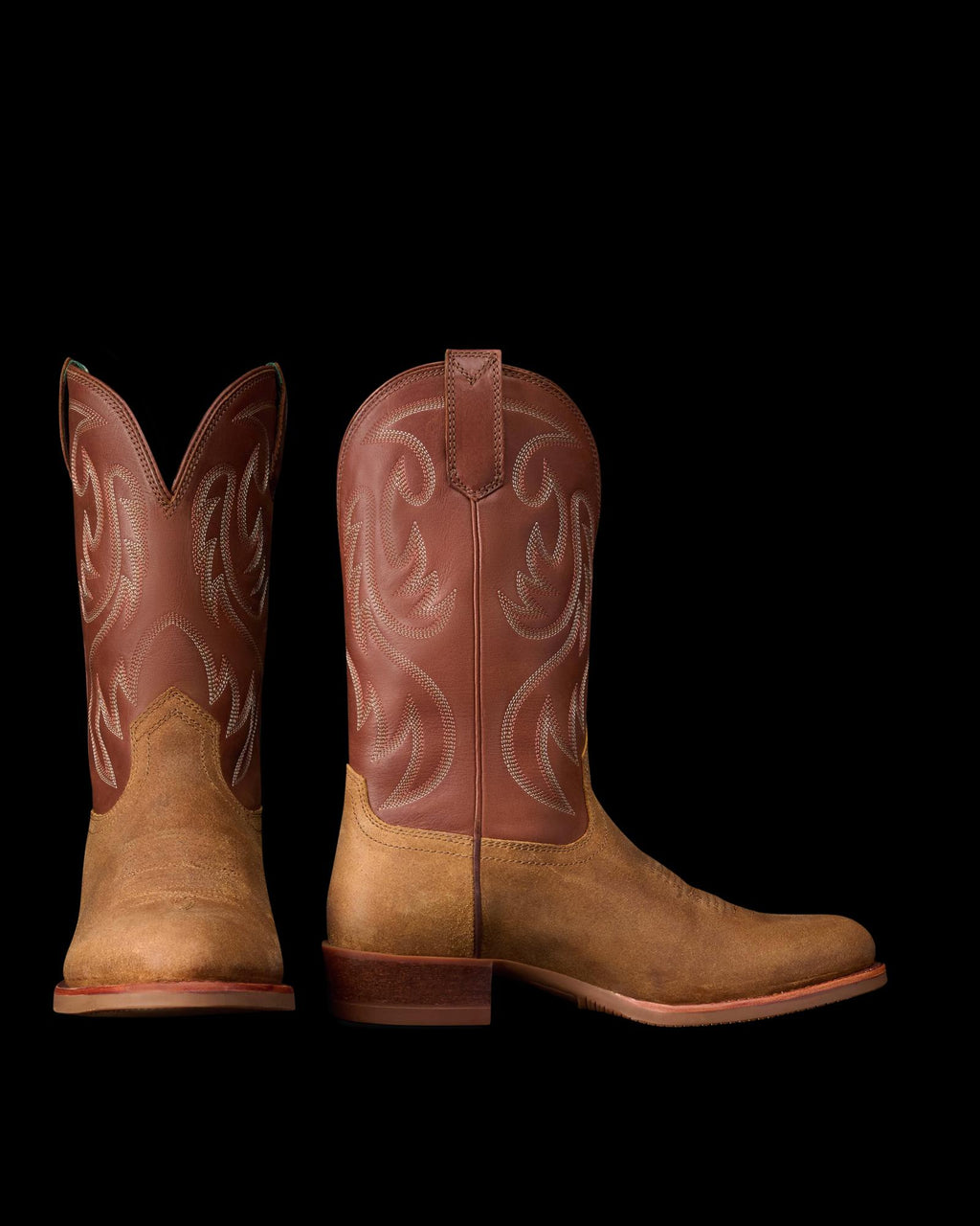 The Buck in Copper Cowhide. Pair of brown western cowboy boots, named The Buck Cowhide, with a light brown foot and darker brown shaft featuring decorative stitching, shown from the front and side on a white background.