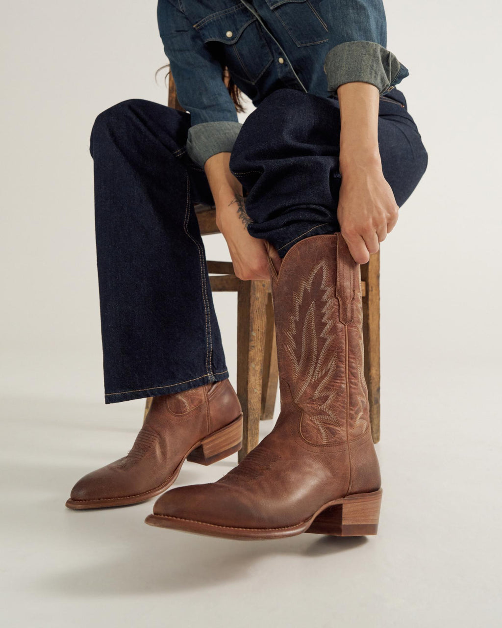 Person in denim outfit sits on a stool, pulling on a pair of brown leather cowboy boots with decorative stitching.