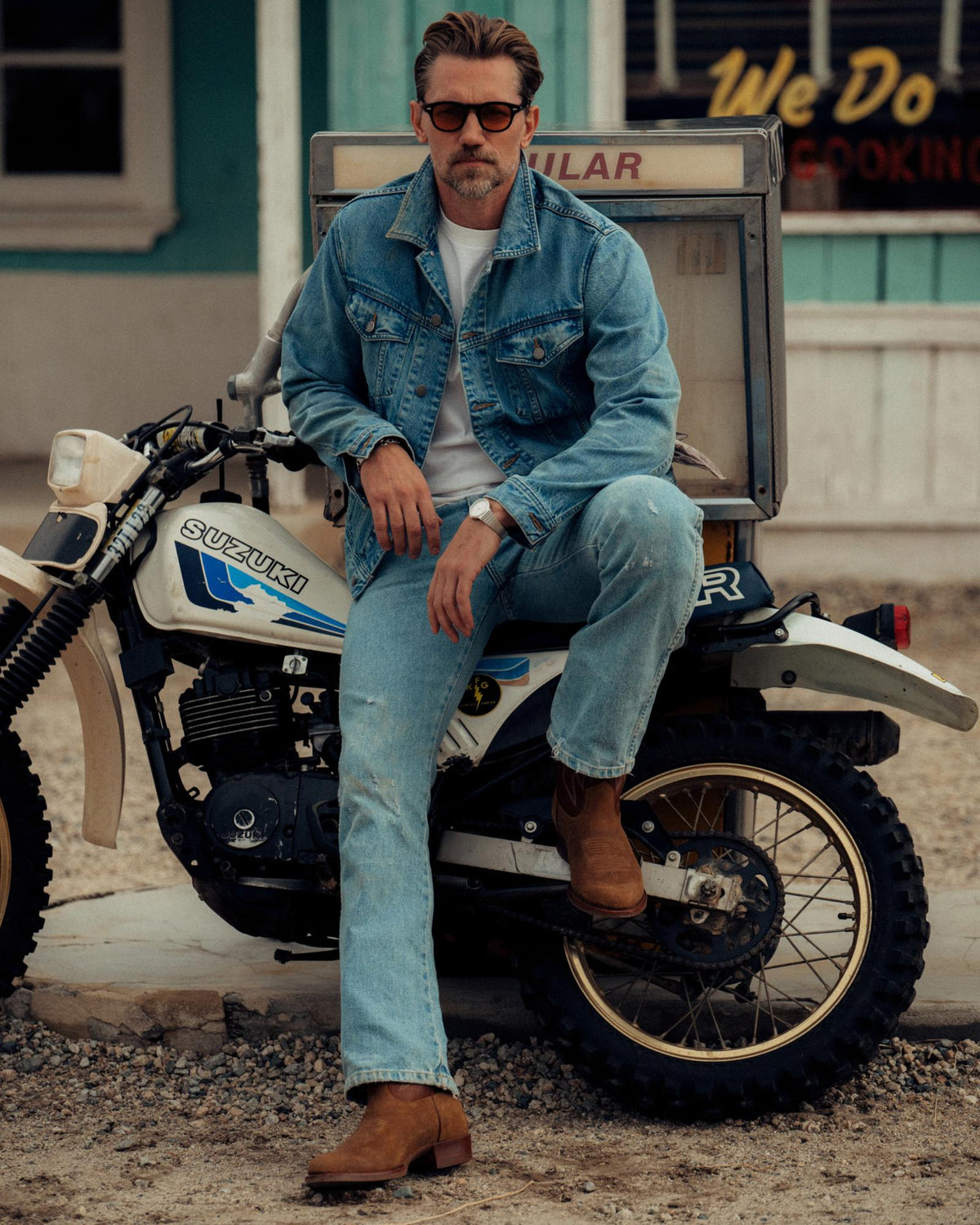 The Buck in Copper Cowhide. A man in sunglasses and denim jacket sits on a Suzuki dirt bike, wearing jeans, brown boots, and The Buck Cowhide gloves, with a building and gravel ground in the background.
