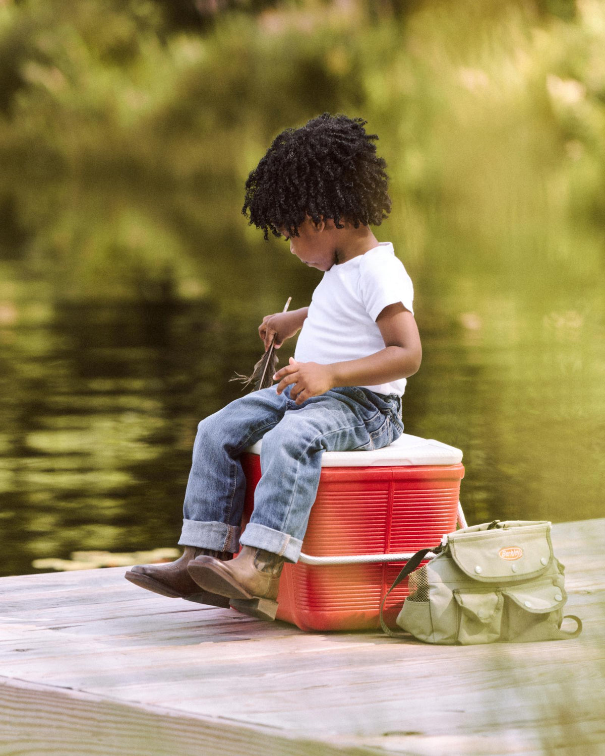 A young child sits on a red cooler by a lake, holding twigs, with a green bag beside them on a wooden dock.