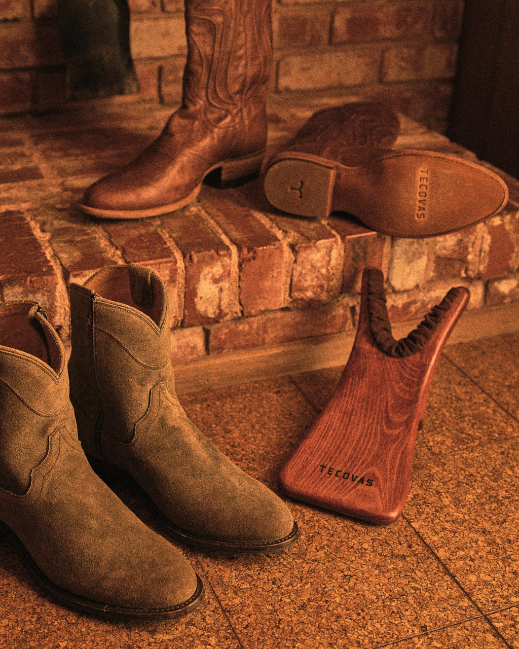 A pair of brown suede cowboy boots, a wooden boot jack, and another pair of leather boots are placed near a brick fireplace.