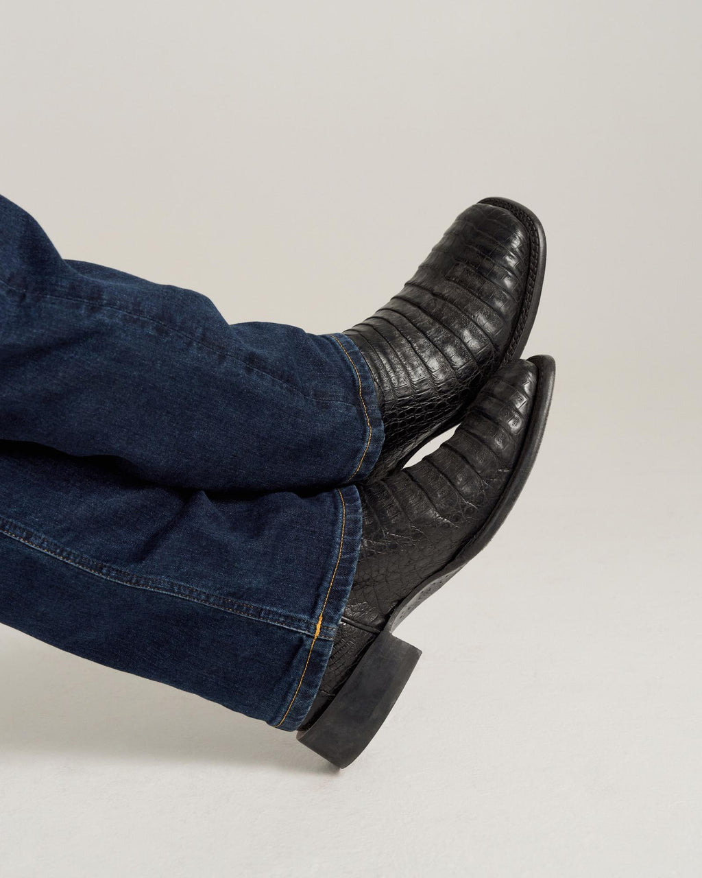 The William in Midnight Caiman. A person wearing dark blue jeans and black textured Midnight Caiman boots is sitting with their legs crossed against a plain background.
