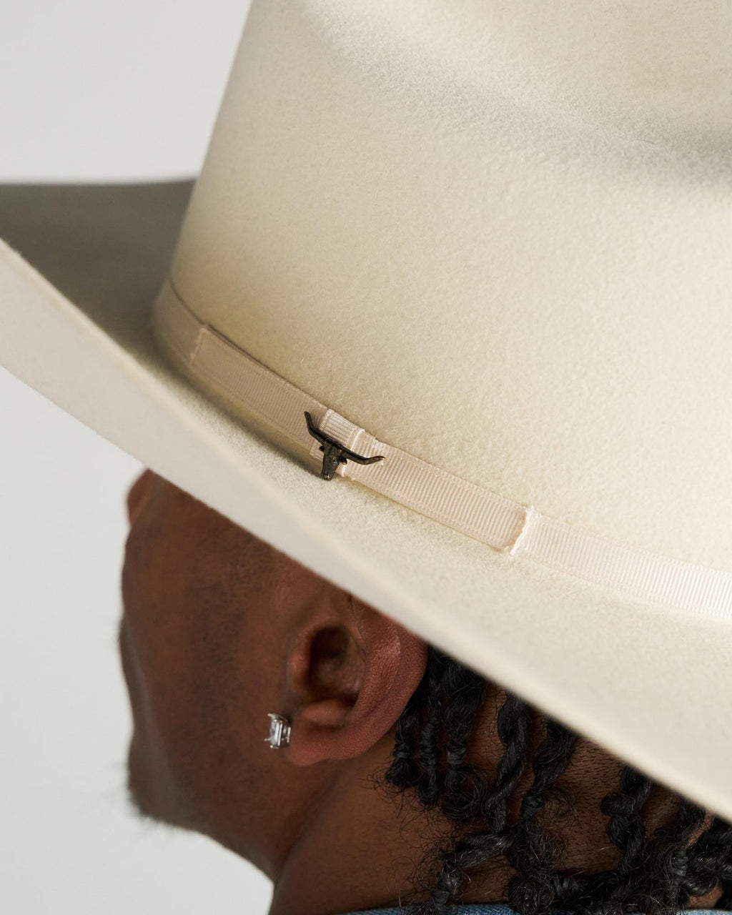 Close-up of a person with braided hair wearing a light-colored wide-brimmed hat, showing the side of their face and an earring.