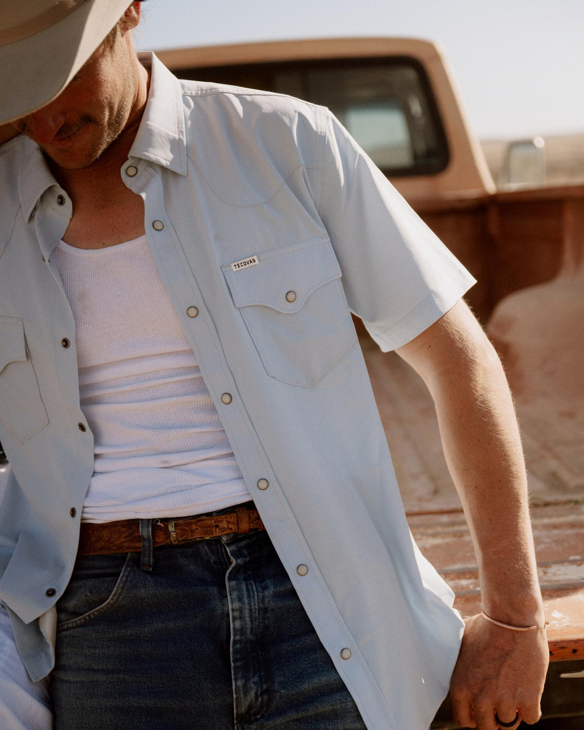 A person wearing a light blue short-sleeve shirt, white tank top, jeans, and a hat stands by an old pickup truck in a sunny outdoor setting.