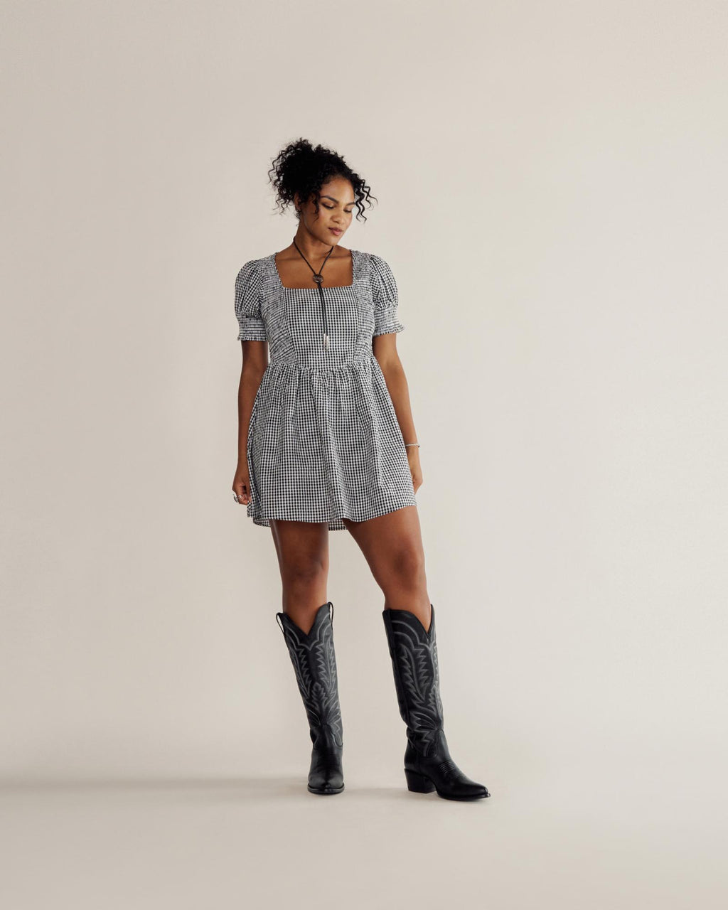 A woman in a black and white checkered dress and black cowboy boots stands against a plain light background, looking slightly downward to her left.