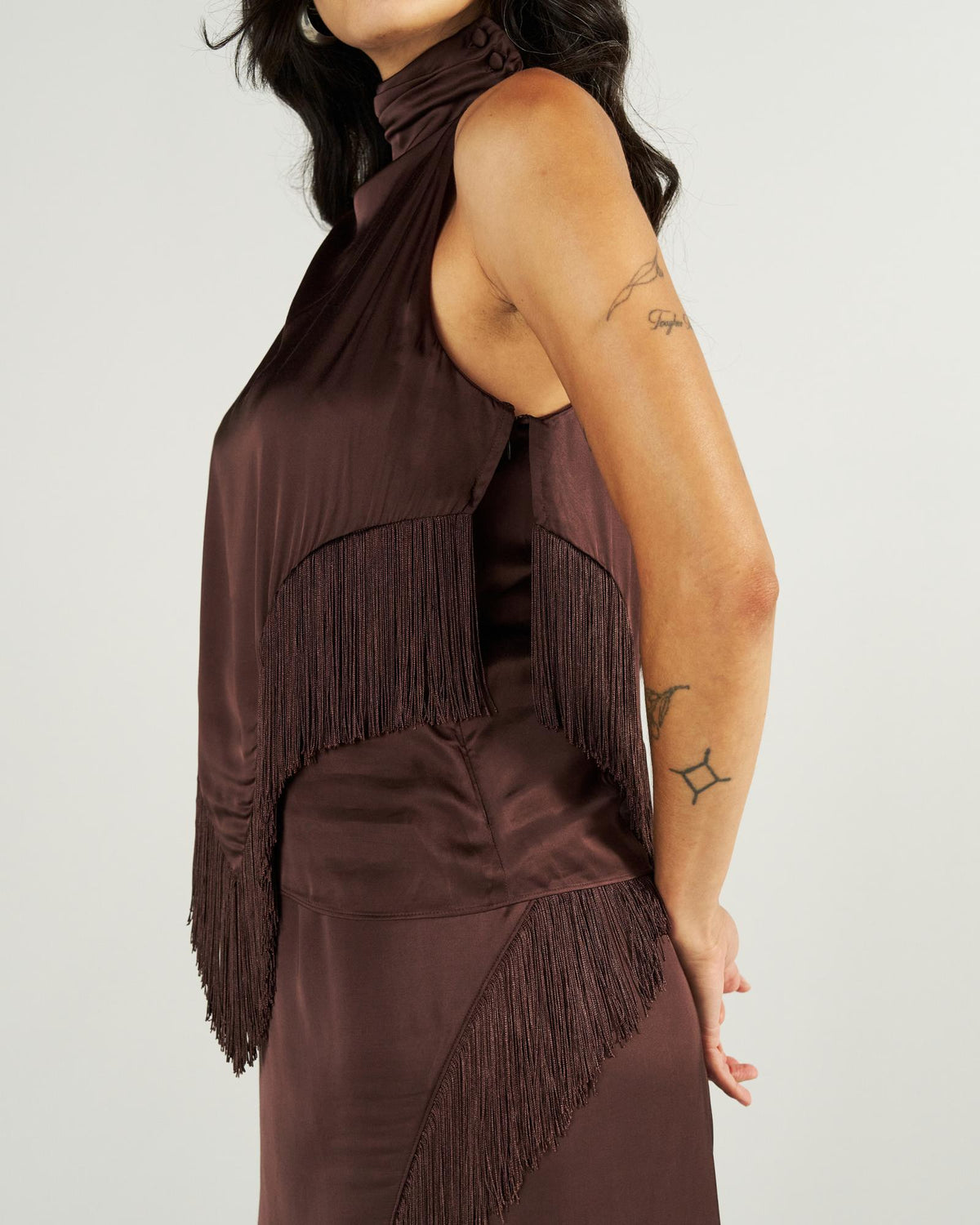 Woman wearing a sleeveless, brown satin dress with fringe details, standing sideways against a plain background. Visible tattoos on her arm.