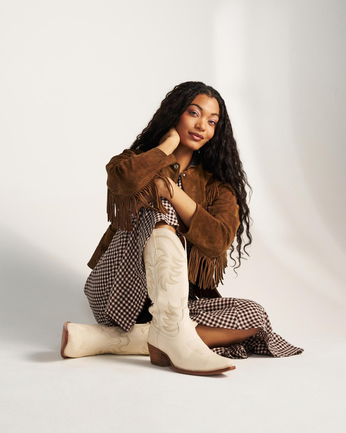 The Annie in Bone Cowhide. Woman sitting on the floor wearing a brown fringed jacket, checkered dress, and The Annie Cowhide white cowboy boots against a plain background.