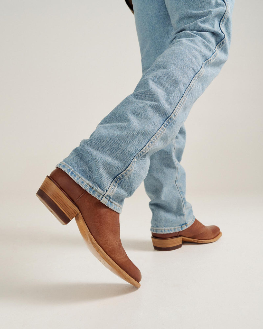 Person wearing light blue jeans and brown leather boots walking, with one foot lifted slightly off the ground against a plain background.