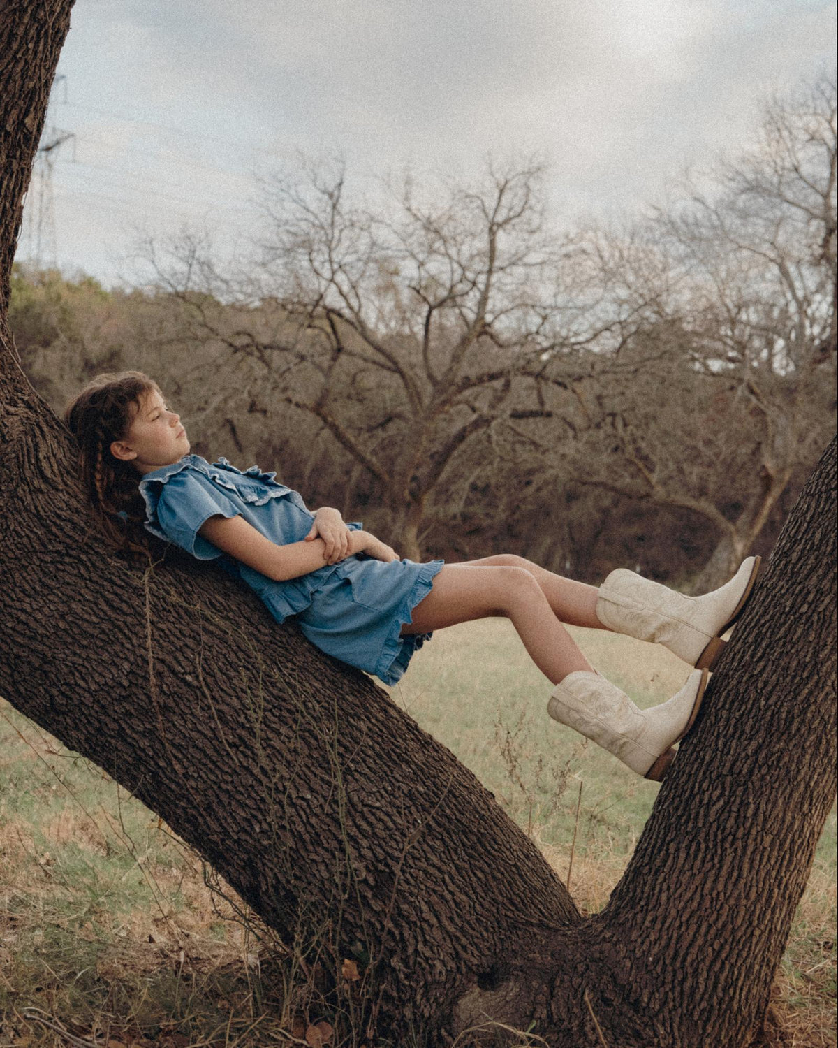 A young girl in a blue dress and white boots lies back on a tree branch in a grassy field, with bare trees in the background.