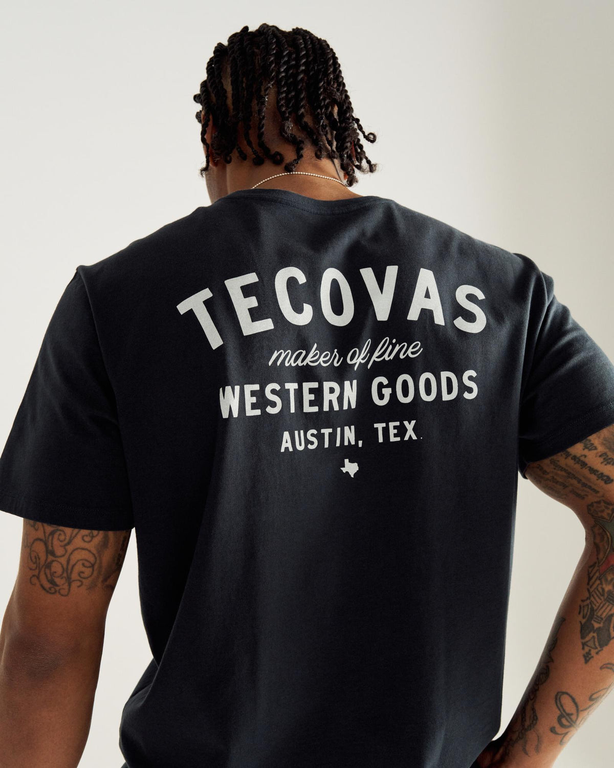 A person with braided hair wears a black T-shirt with white text on the back reading "Tecovas maker of fine Western Goods, Austin, Tex.