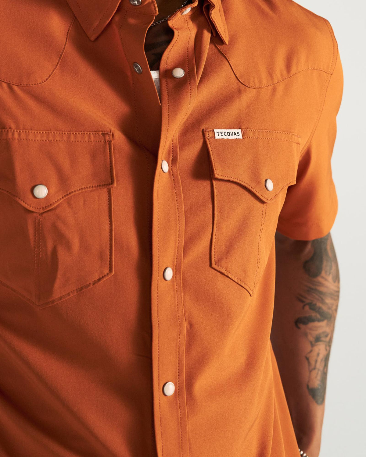Person wearing an orange short-sleeve button-up shirt with two chest pockets and visible tattoos on their arm.