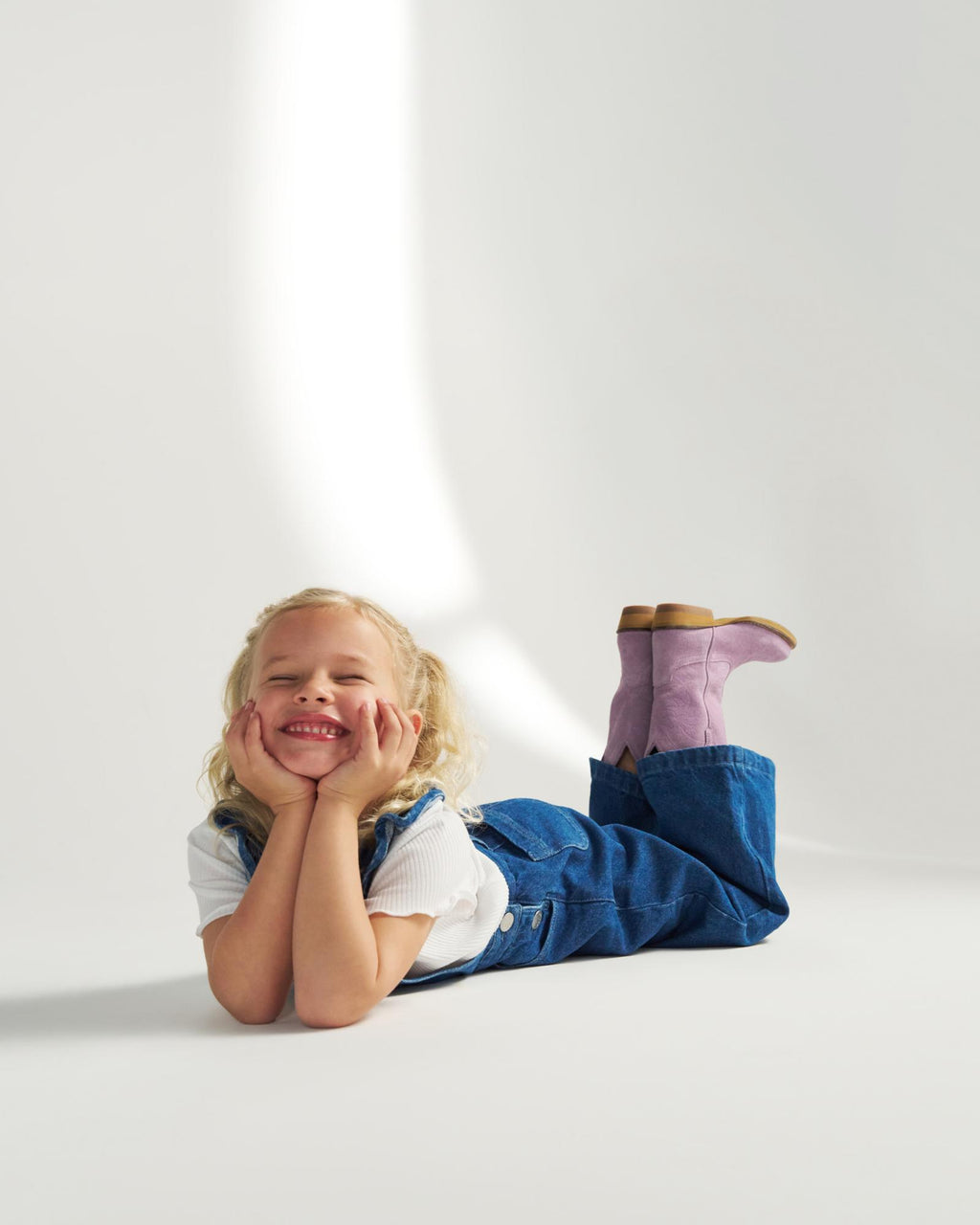 A young child with blonde hair, wearing a white shirt, blue overalls, and pink boots, lies on the floor smiling with hands under their chin.