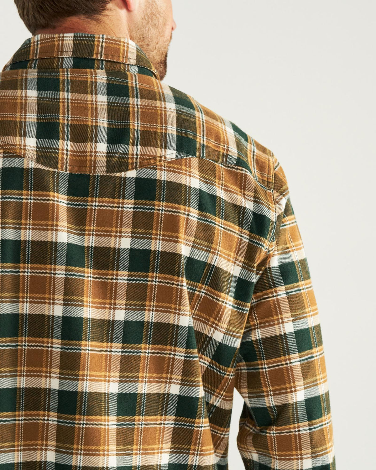 A man wearing a green, brown, and white plaid flannel shirt is shown from behind, with the focus on the shirt’s pattern and texture.