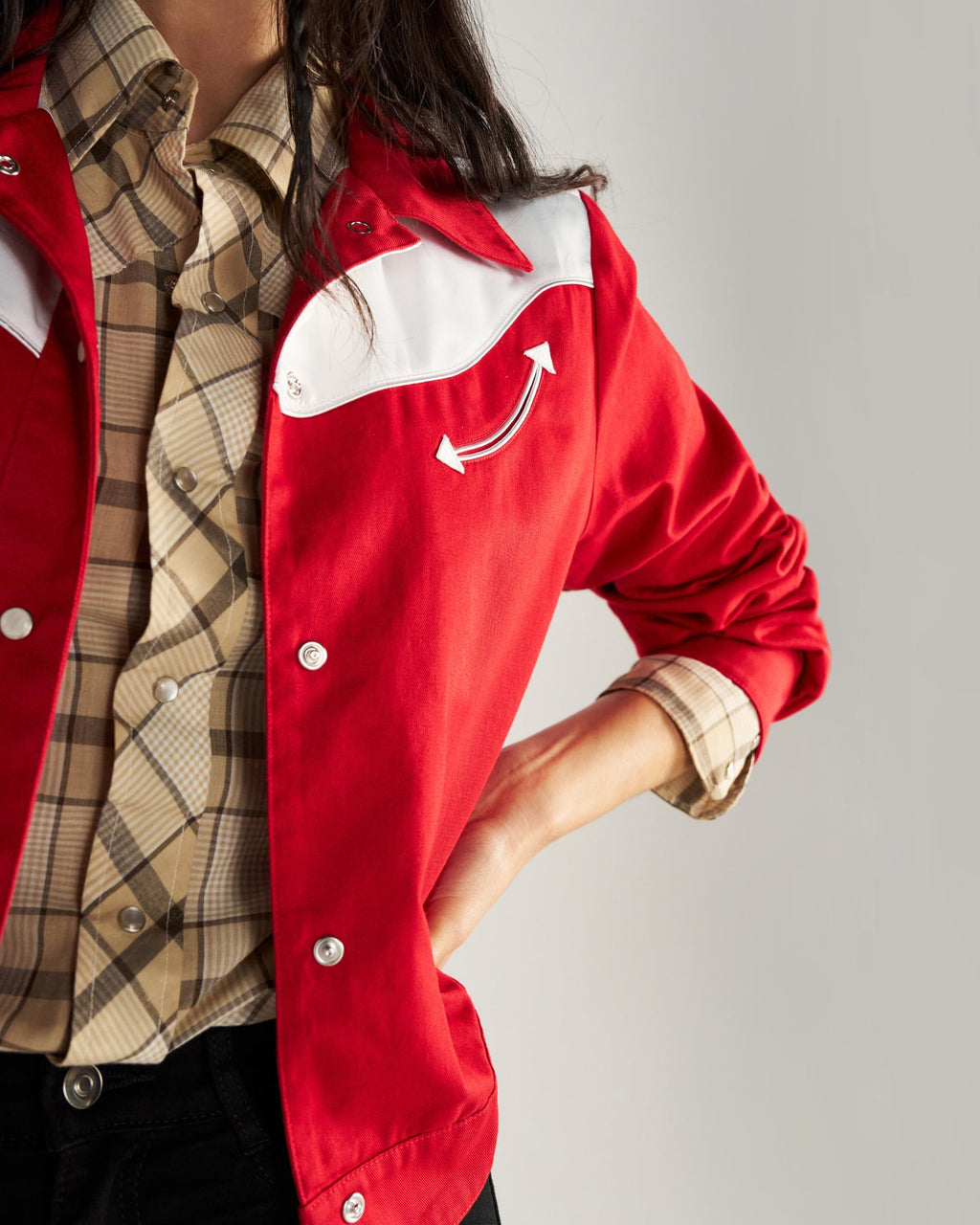 A person stands with one hand on their hip, wearing a red jacket with white detailing over a plaid shirt and black pants.