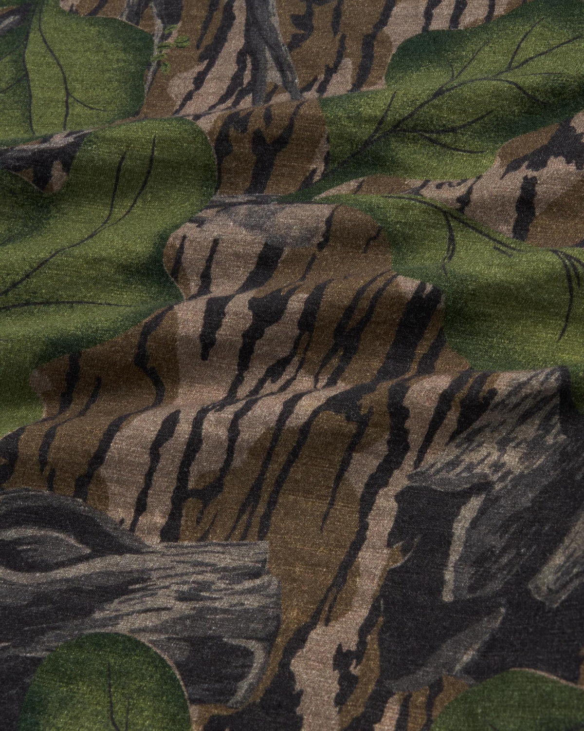 A close-up view of camouflage fabric with a pattern resembling tree bark, branches, and green leaves.