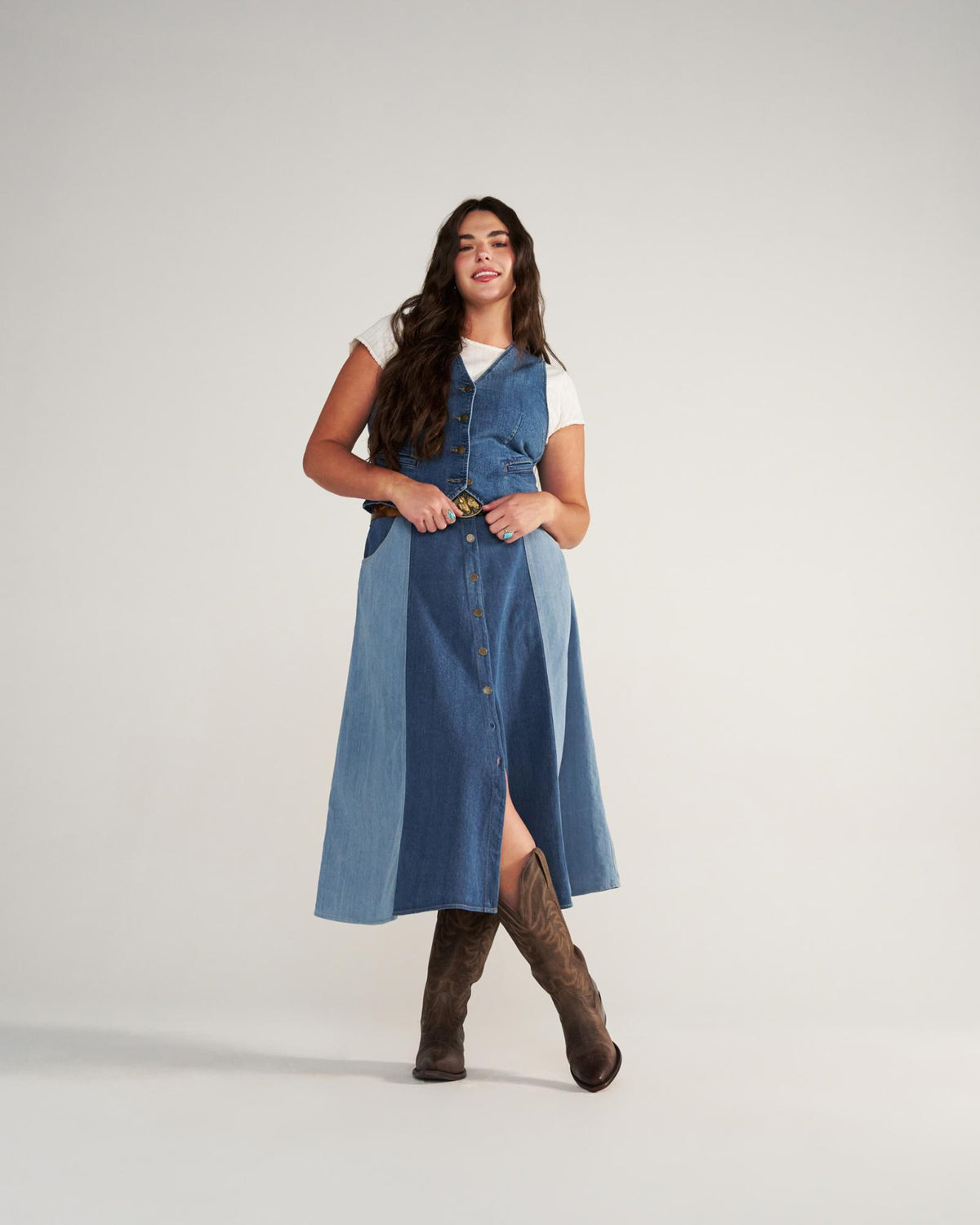 The Annie Wide Calf in Cafe Goat. A woman stands in a studio wearing The Annie Wide Calf Goat denim button-front dress over a white t-shirt, a belt, and brown cowboy boots.