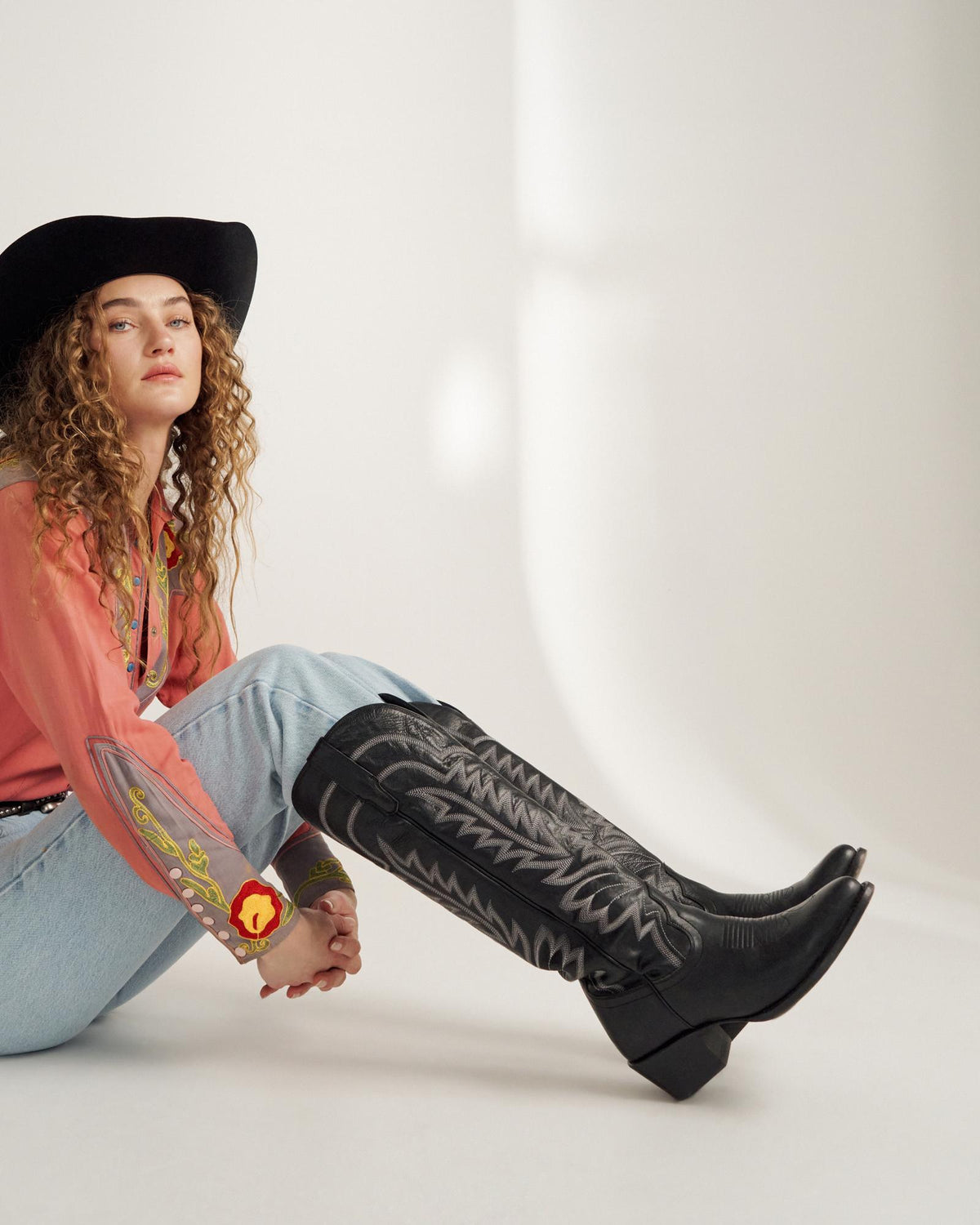 A person in a black cowboy hat, coral western shirt, light blue jeans, and black cowboy boots sits on the floor against a white background.