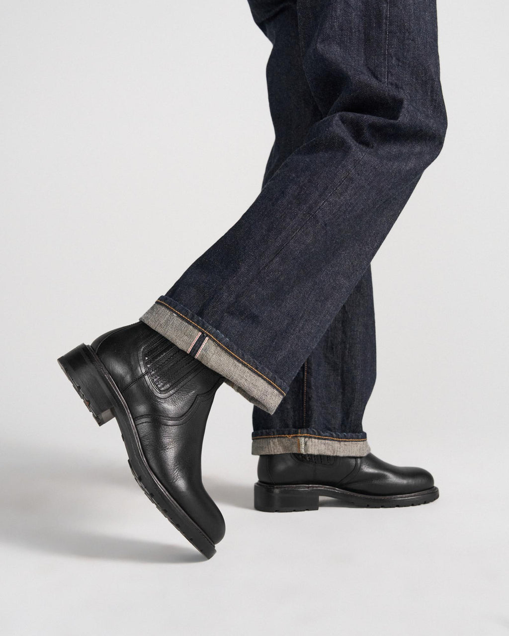 The Chelsea in Midnight Cowhide. Person wearing dark cuffed jeans and black leather boots, standing with one foot slightly raised against a plain background—the Midnight Cowhide boots add a refined edge to this timeless look.