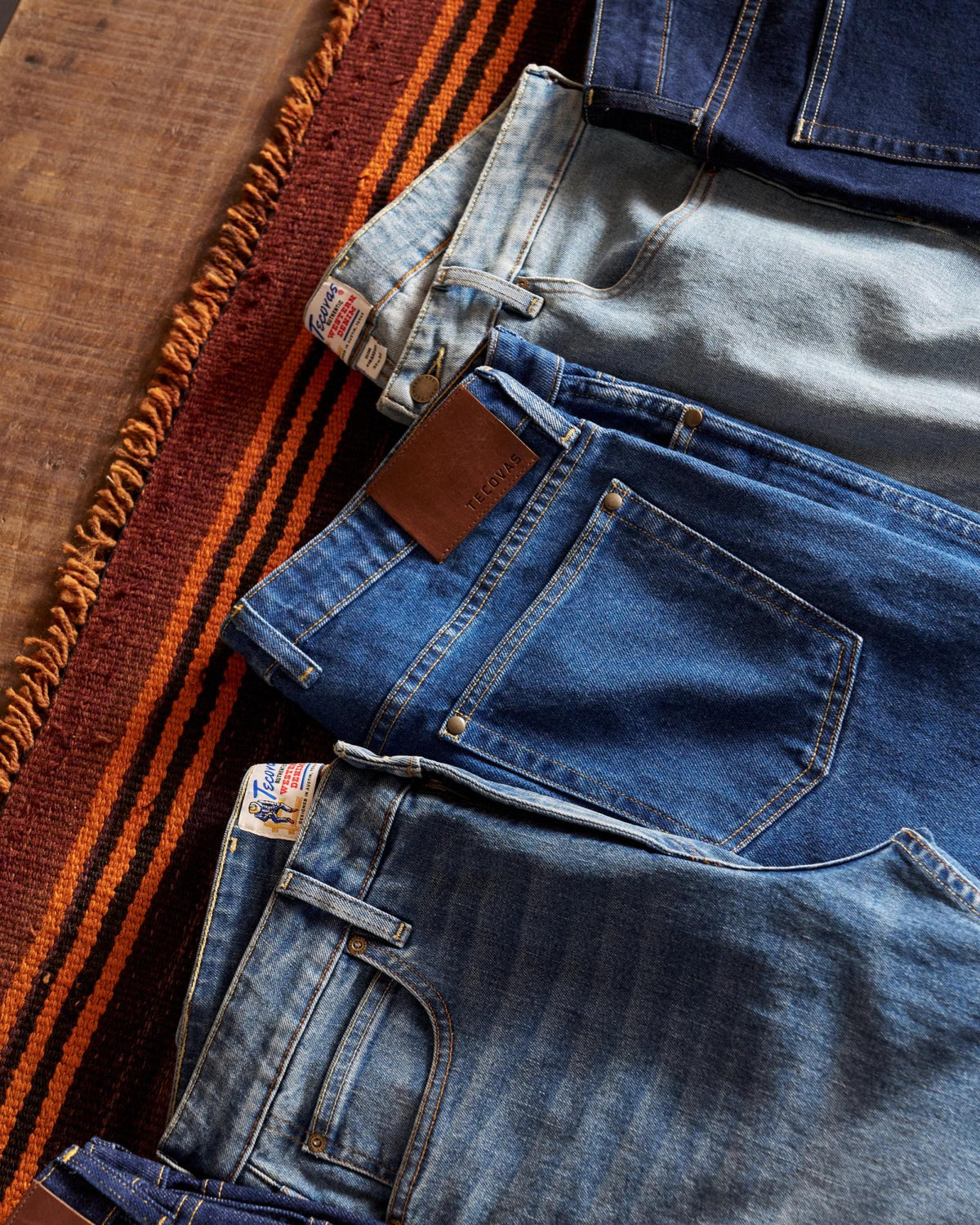Four pairs of blue jeans in different washes are laid out side by side on a woven fabric with orange and black stripes, next to a wooden surface.