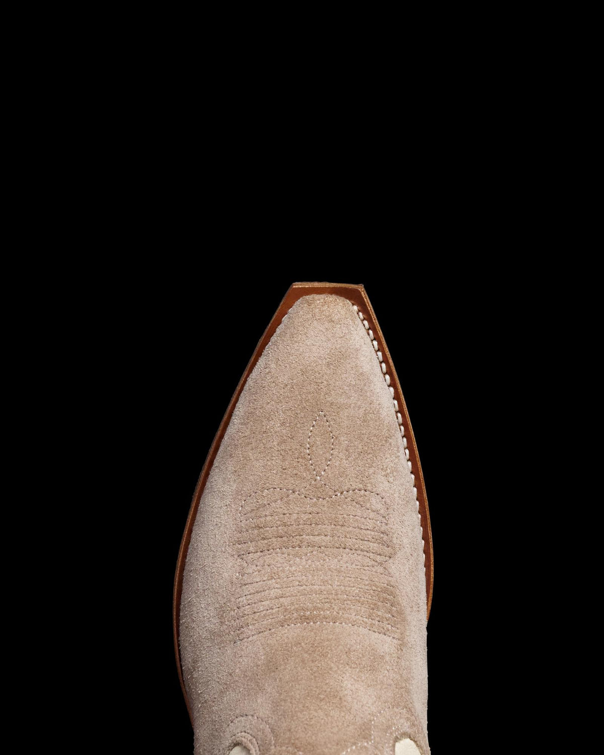 Close-up of the toe of a beige suede cowboy boot with stitch detailing, isolated on a black background.