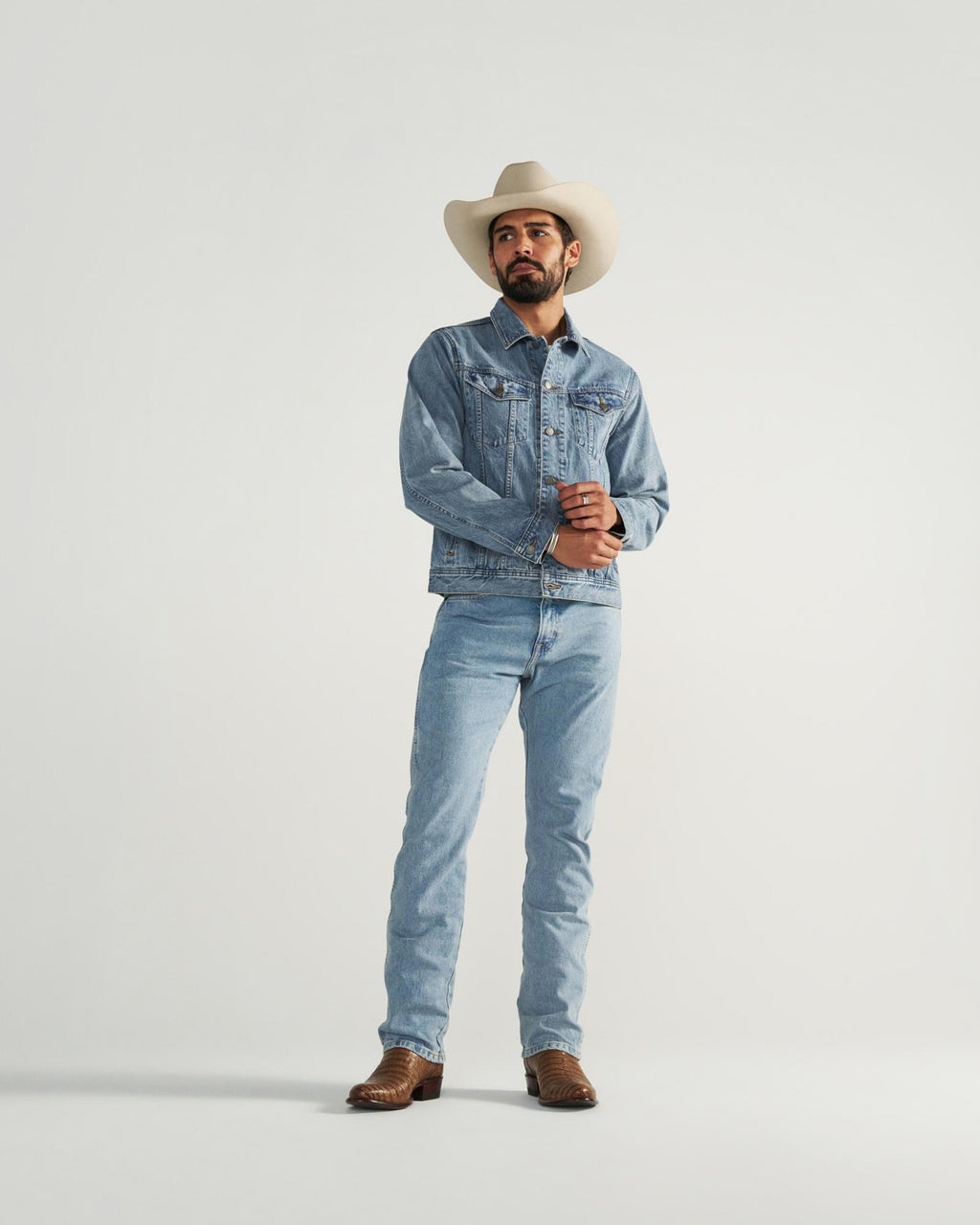A man wearing a white cowboy hat, denim jacket, denim jeans, and brown cowboy boots stands against a plain light background.