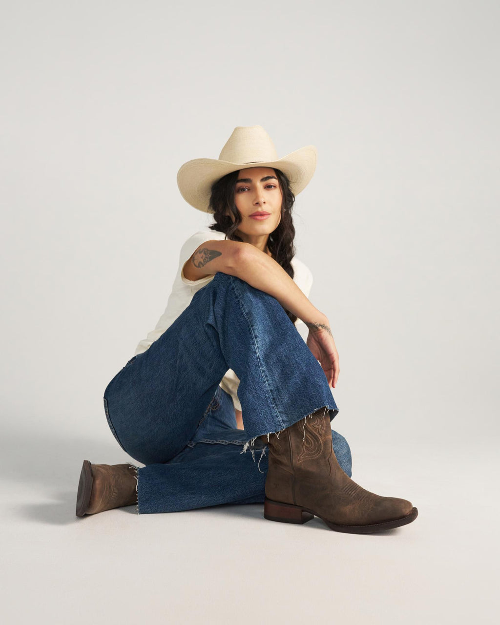 Person wearing a cowboy hat, white shirt, blue jeans, and brown boots sits on the floor against a plain background, reminiscent of a relaxed afternoon at The Tess Goat Cafe.