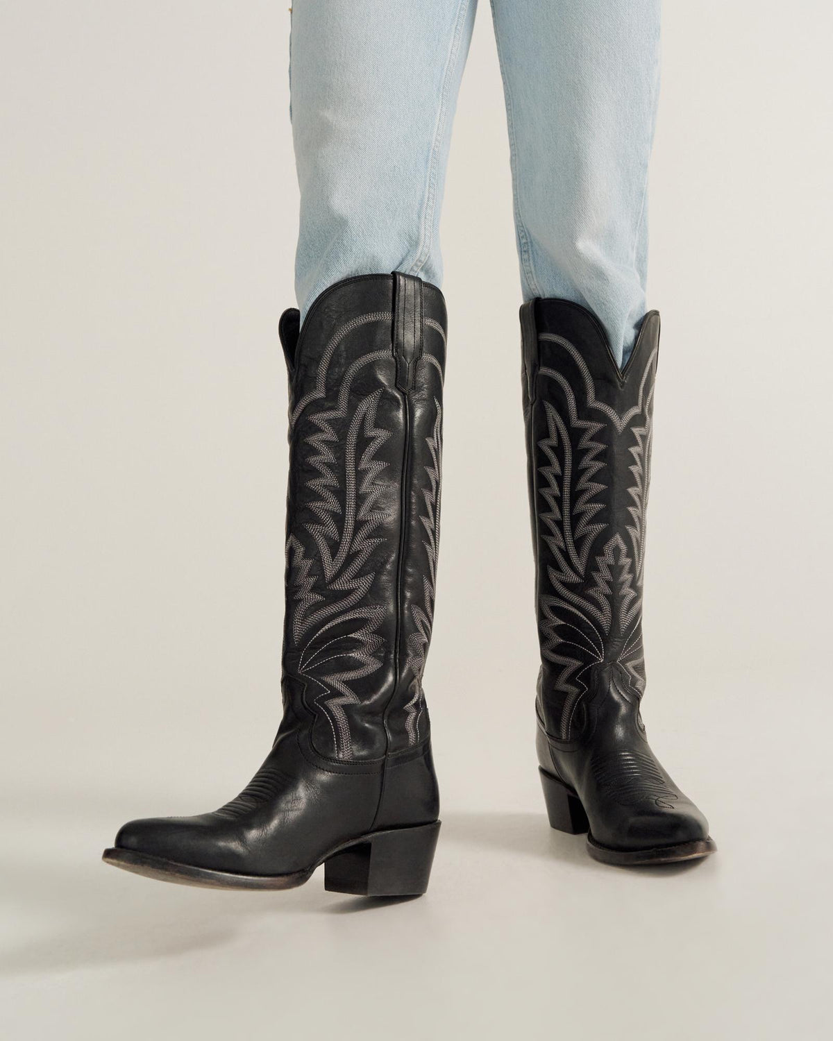 A person wearing light blue jeans and black western cowboy boots stands on a light-colored surface.